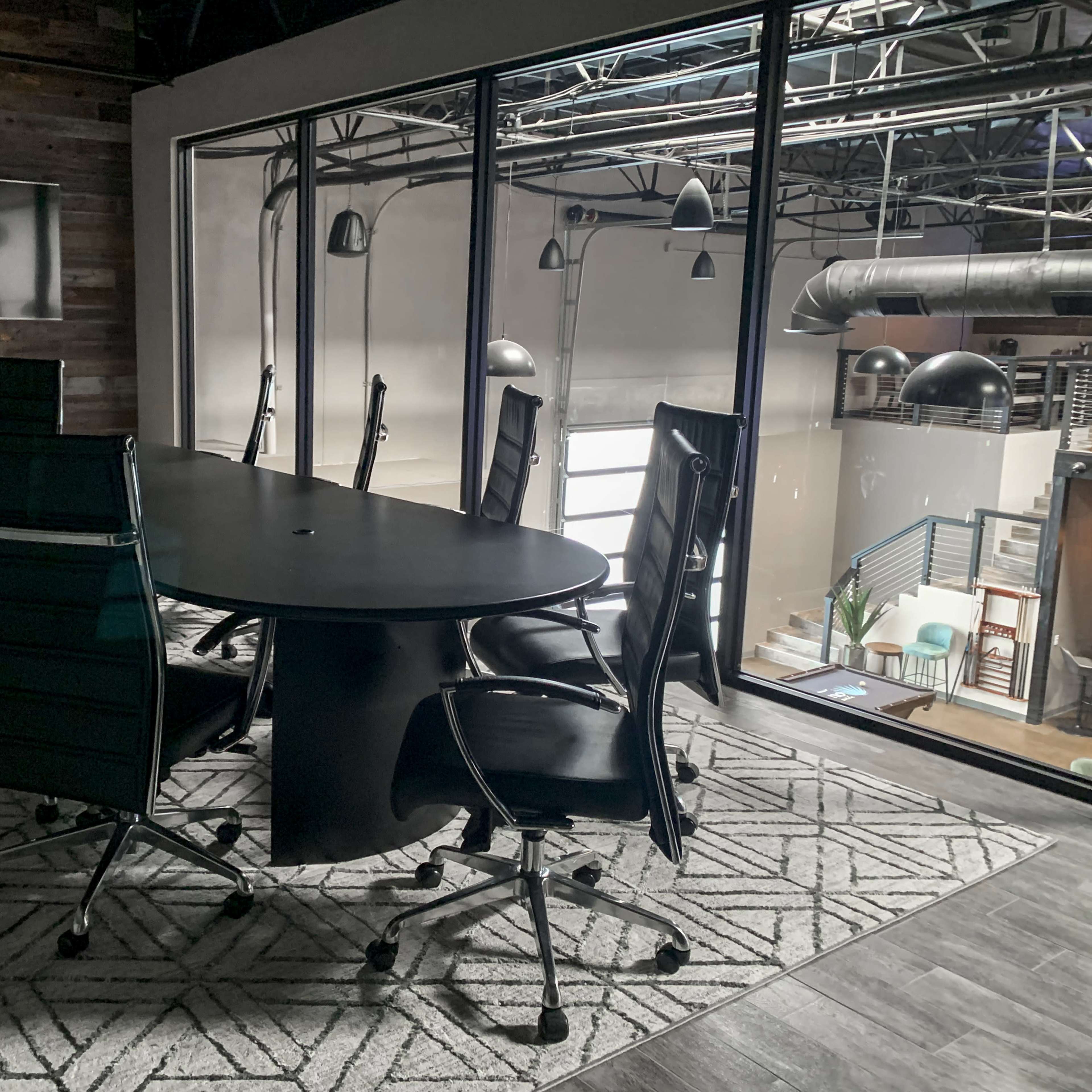 A modern conference room features a large oval table surrounded by eight ergonomic chairs, with large windows overlooking an industrial space.