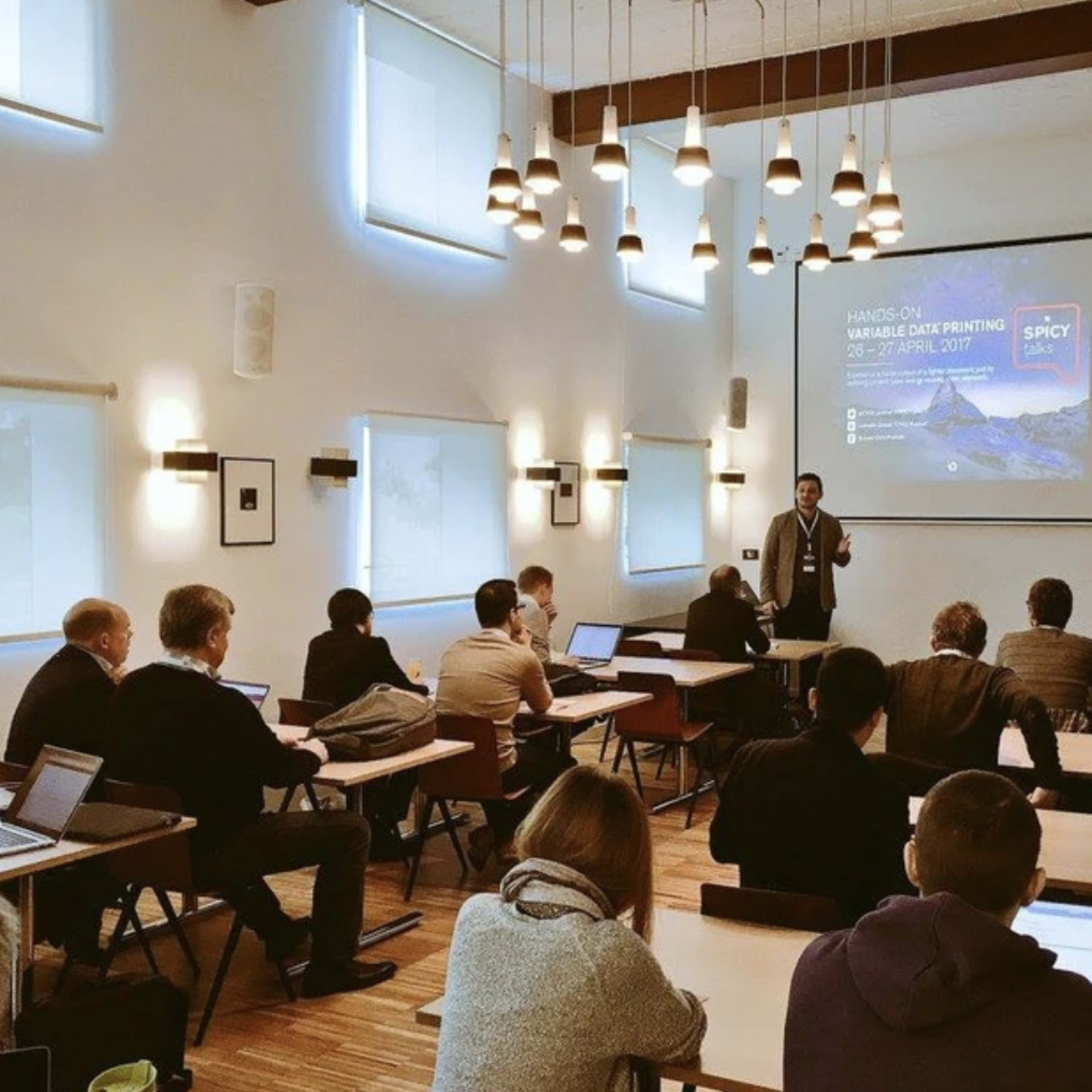 A speaker presents to an audience seated at tables in a classroom setting, with laptops open and a projector displaying information on a screen.
