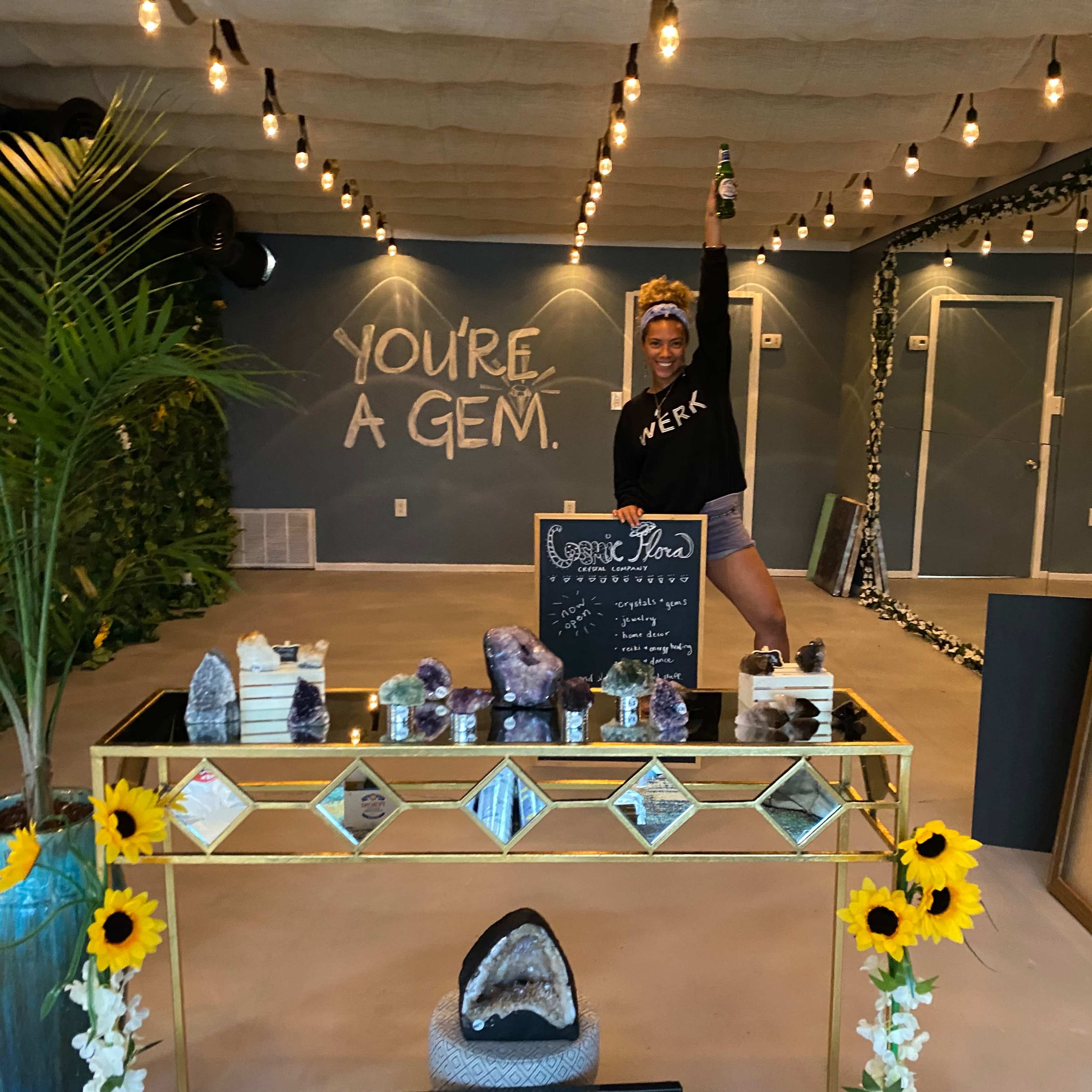 A person stands with one arm raised behind a table displaying various crystals and decorative items in a brightly lit space.
