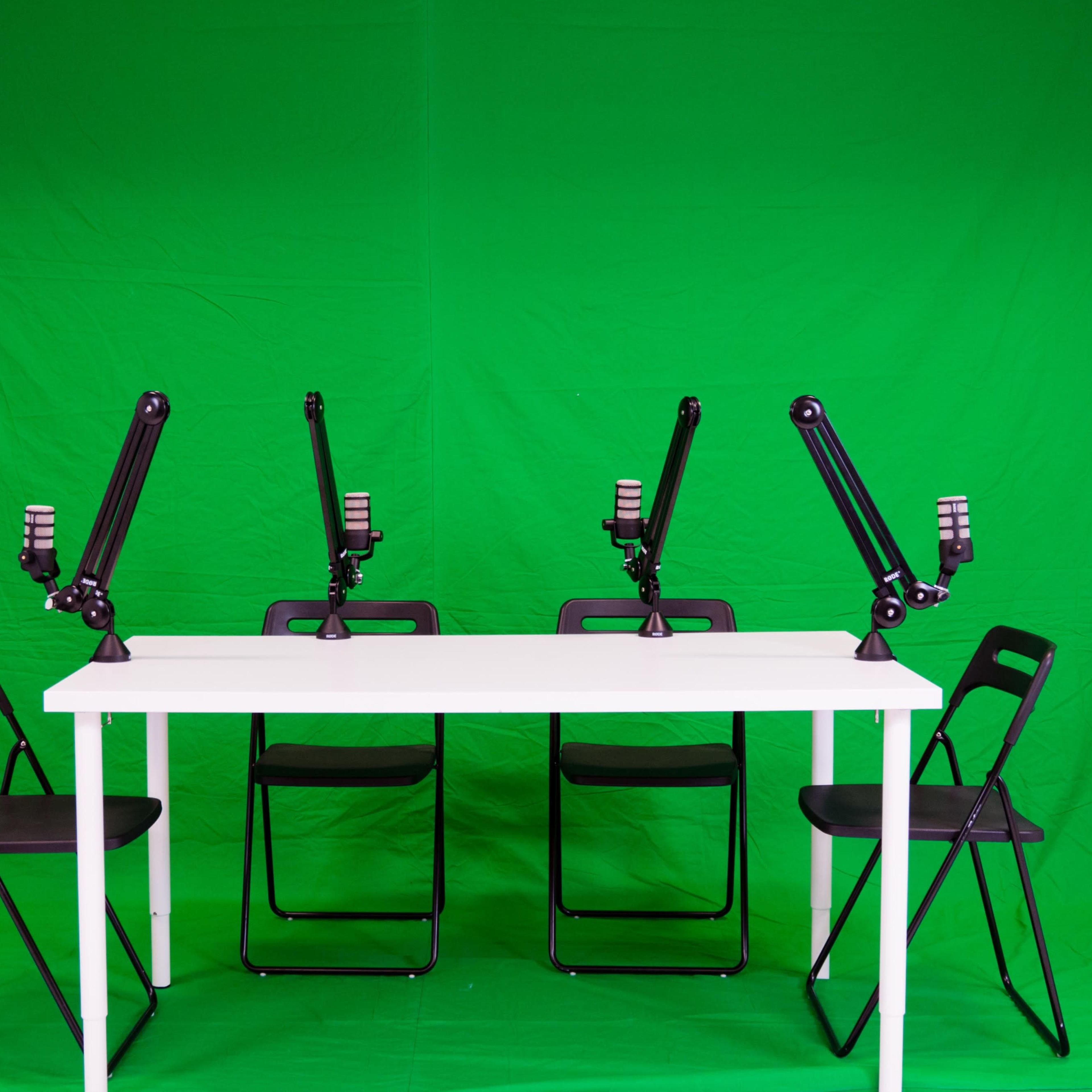A white table is set up in front of a green backdrop with four black chairs and microphone stands positioned evenly along the table.