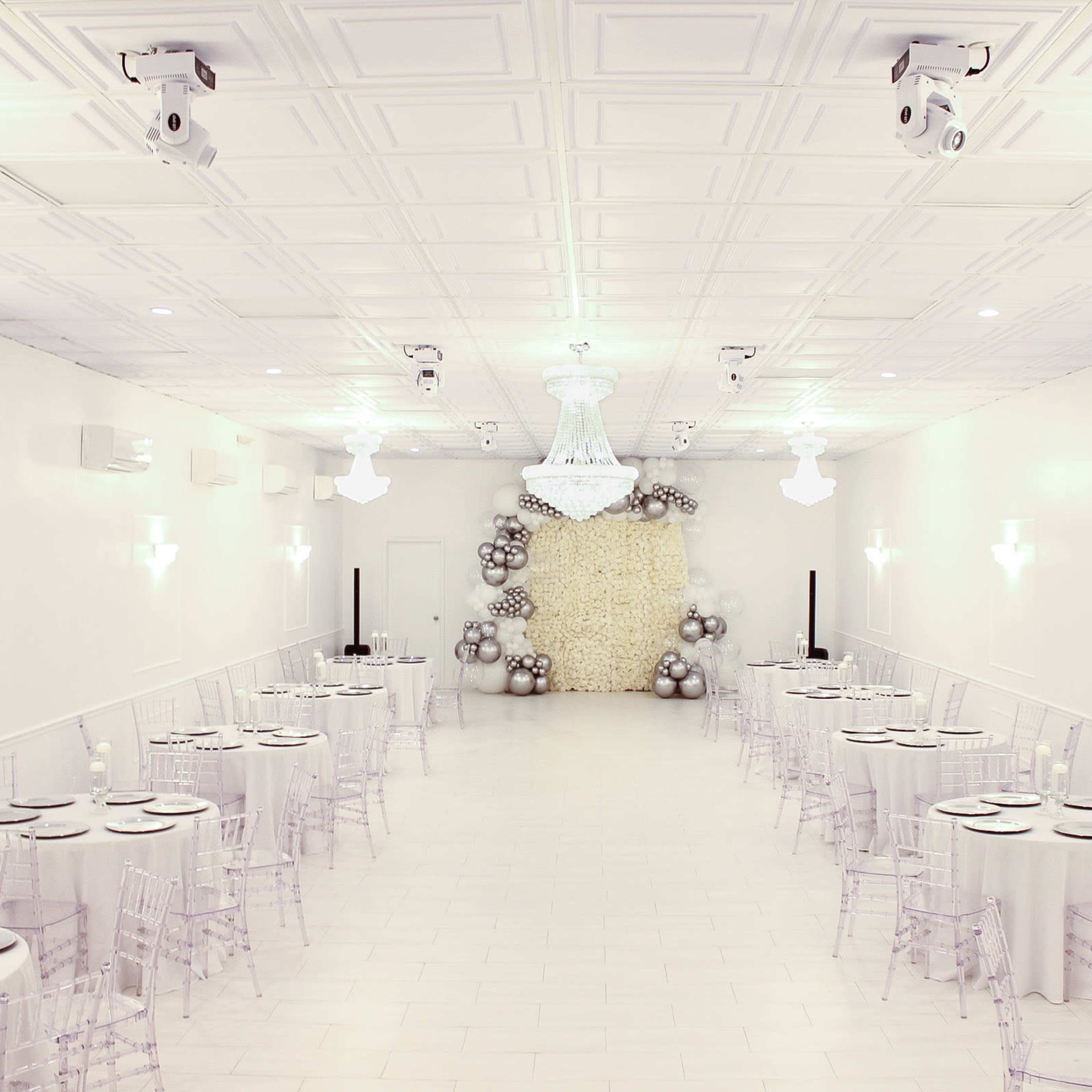 A spacious, elegantly set dining hall with round tables covered in light-colored tablecloths, transparent chairs, and a decorative wall featuring a cluster of balloons.
