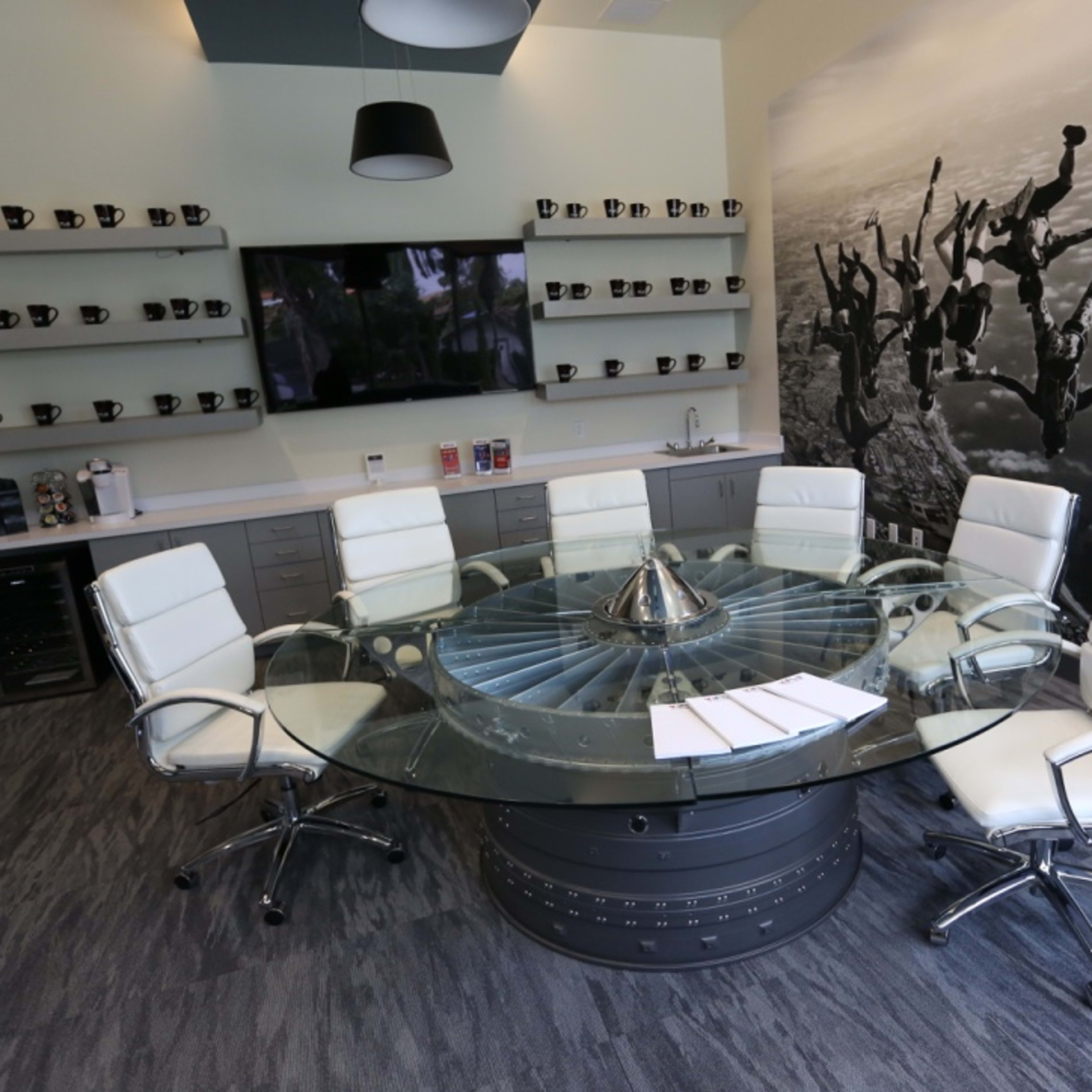 A modern conference room features a glass circular table surrounded by white leather chairs, with a wall mural of people skydiving and shelves displaying coffee mugs.