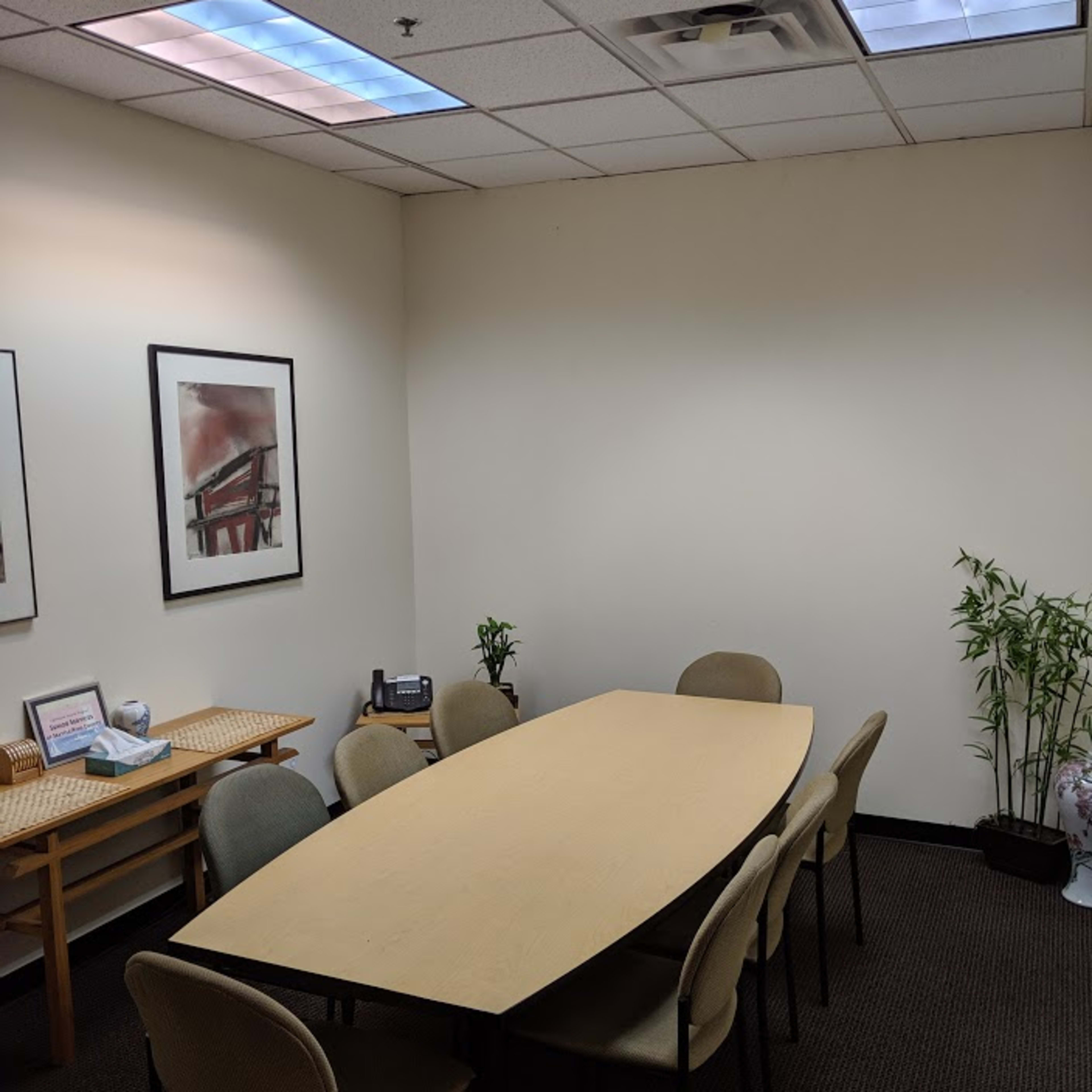 A conference room features a long table surrounded by chairs, with framed artwork on the walls and plants in the corners.