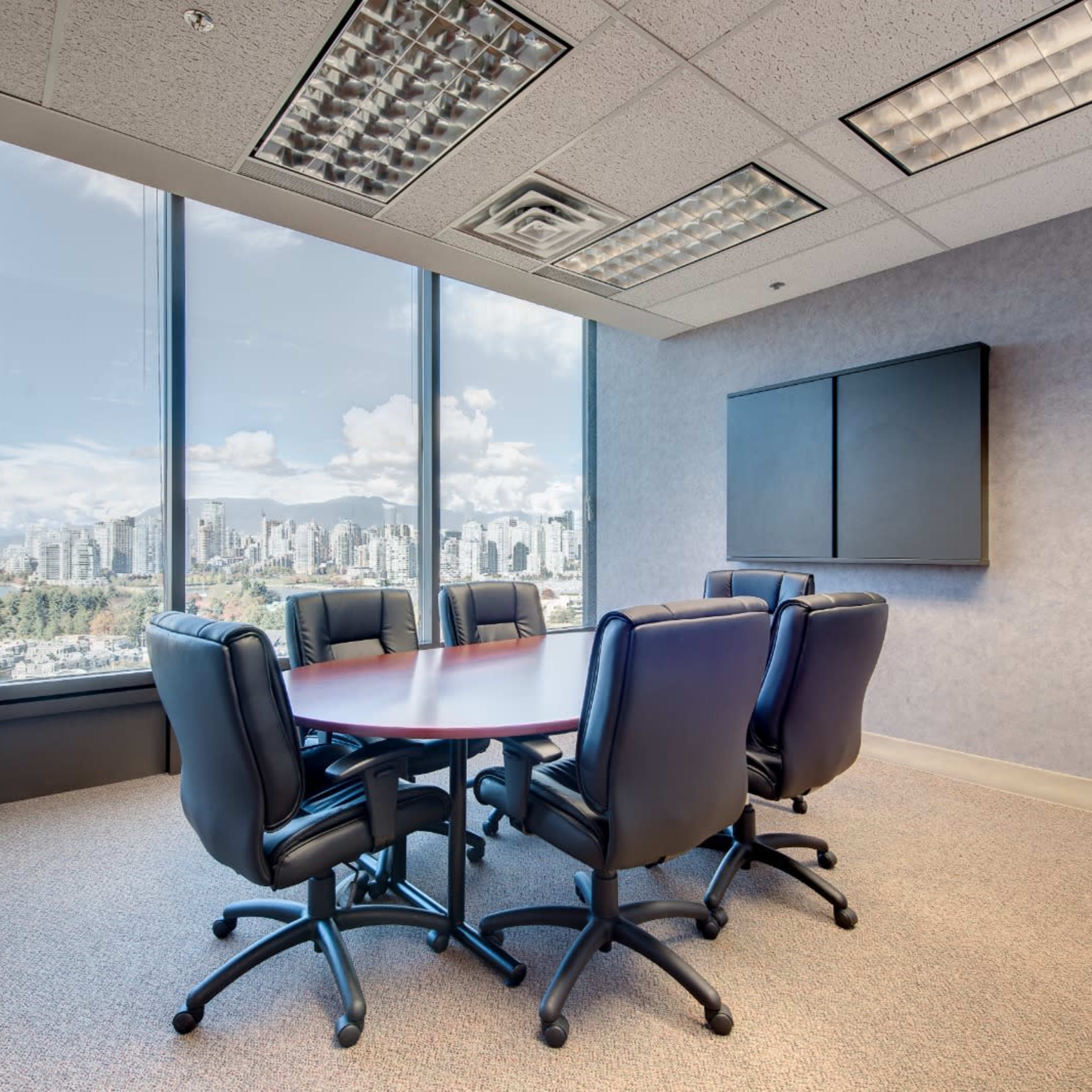 A conference room with a round table surrounded by six black office chairs, featuring large windows that overlook a city skyline.