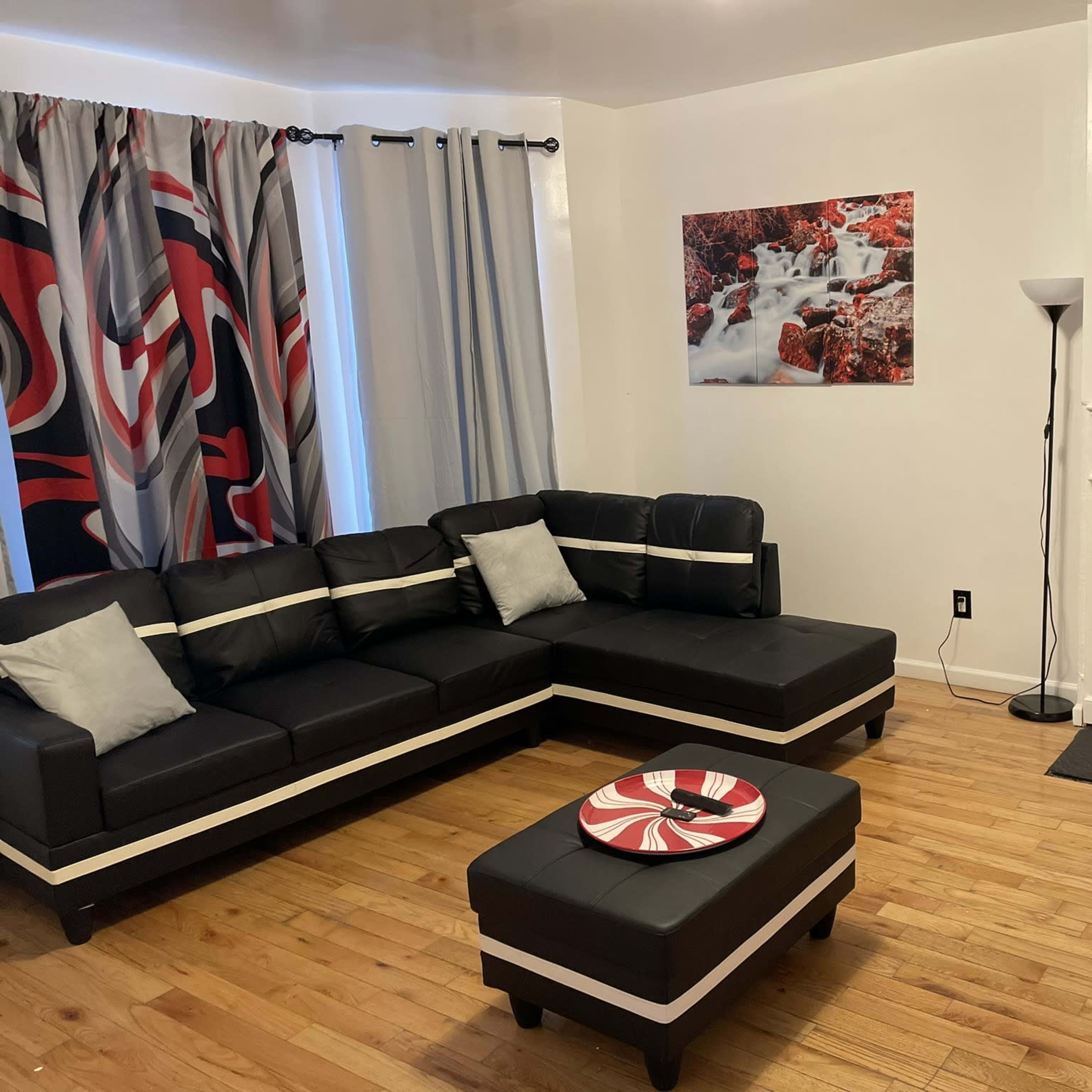 The living room features a black sectional couch, a round coffee table with a decorative plate, a red fireplace, and a large photograph of a water scene on the wall, with curtains displaying a bold pattern.