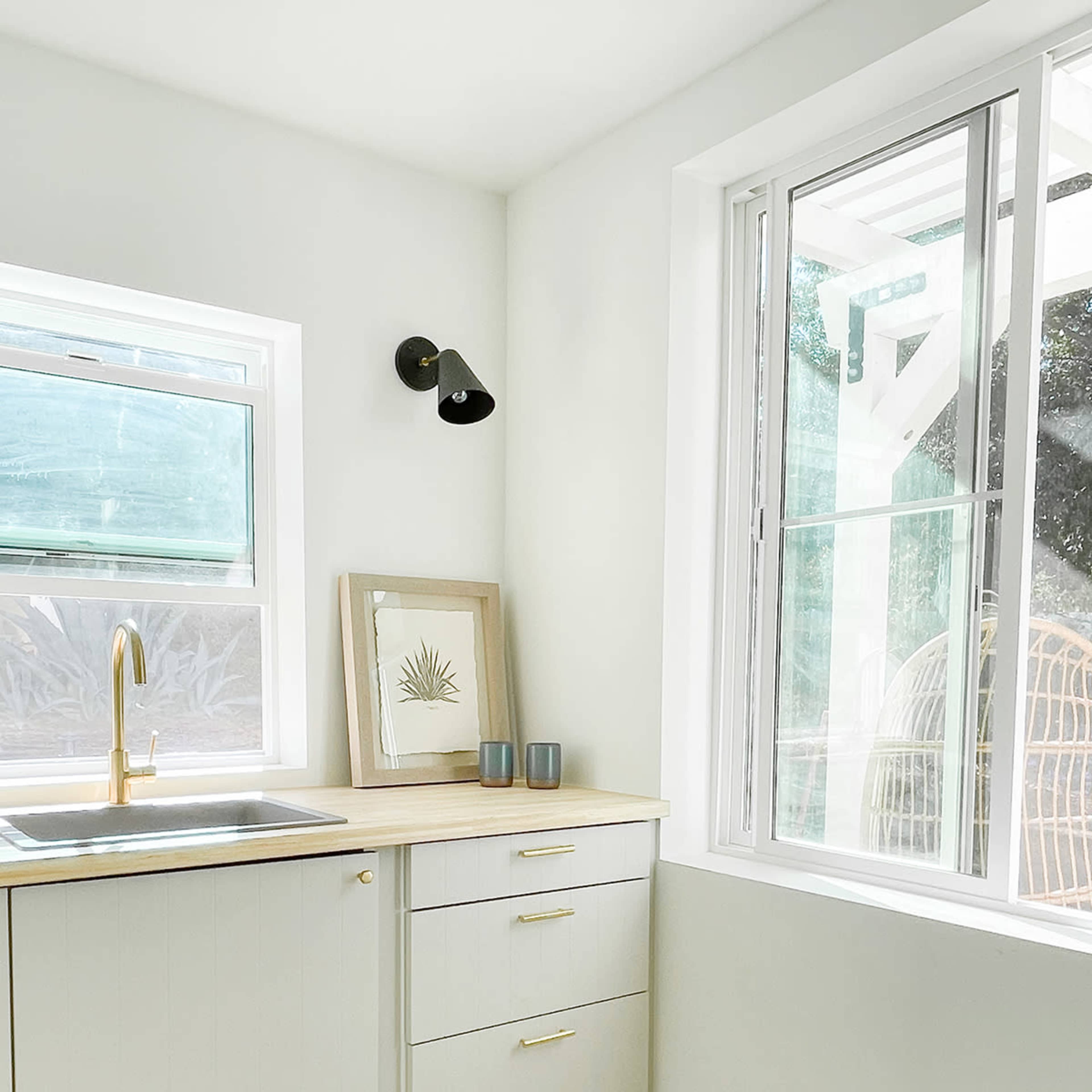 A modern kitchen corner with a sink, wooden countertop, framed artwork, and natural light coming through a window.