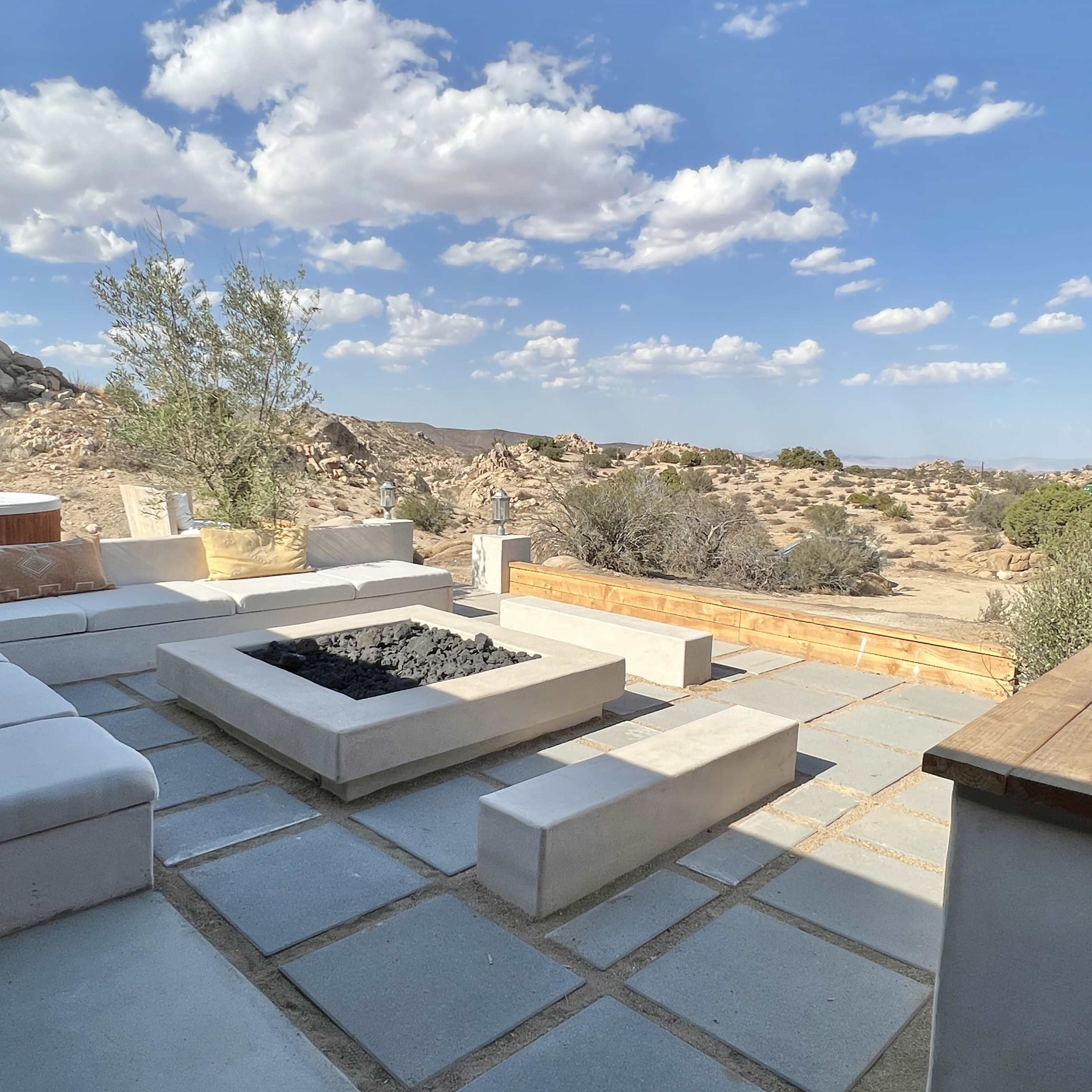A modern outdoor seating area with a fire pit surrounded by stone slabs, set against a rocky desert landscape under a partly cloudy sky.