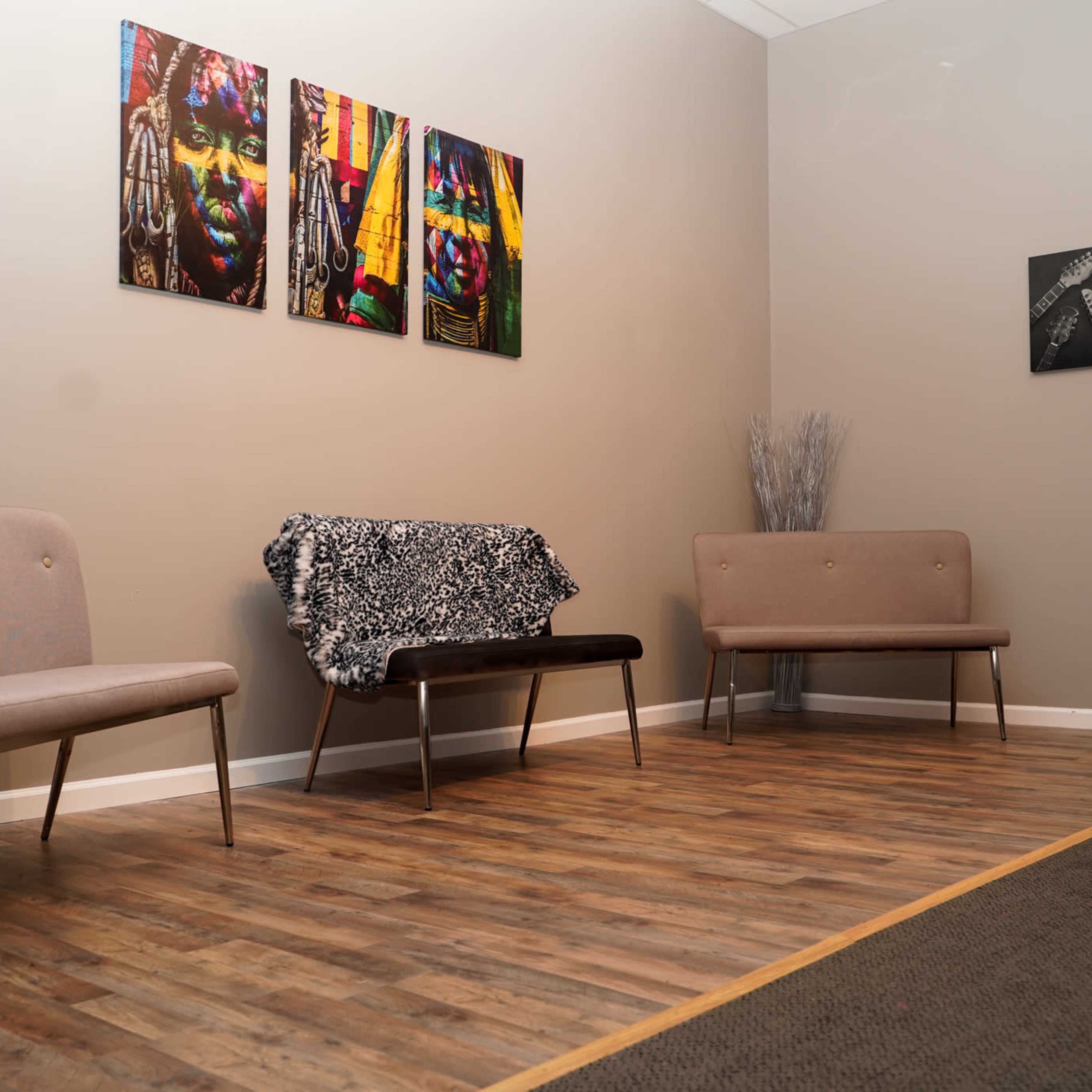A waiting area with two minimalist chairs, a black bench, and three colorful paintings on the wall, along with a decorative element in the corner.