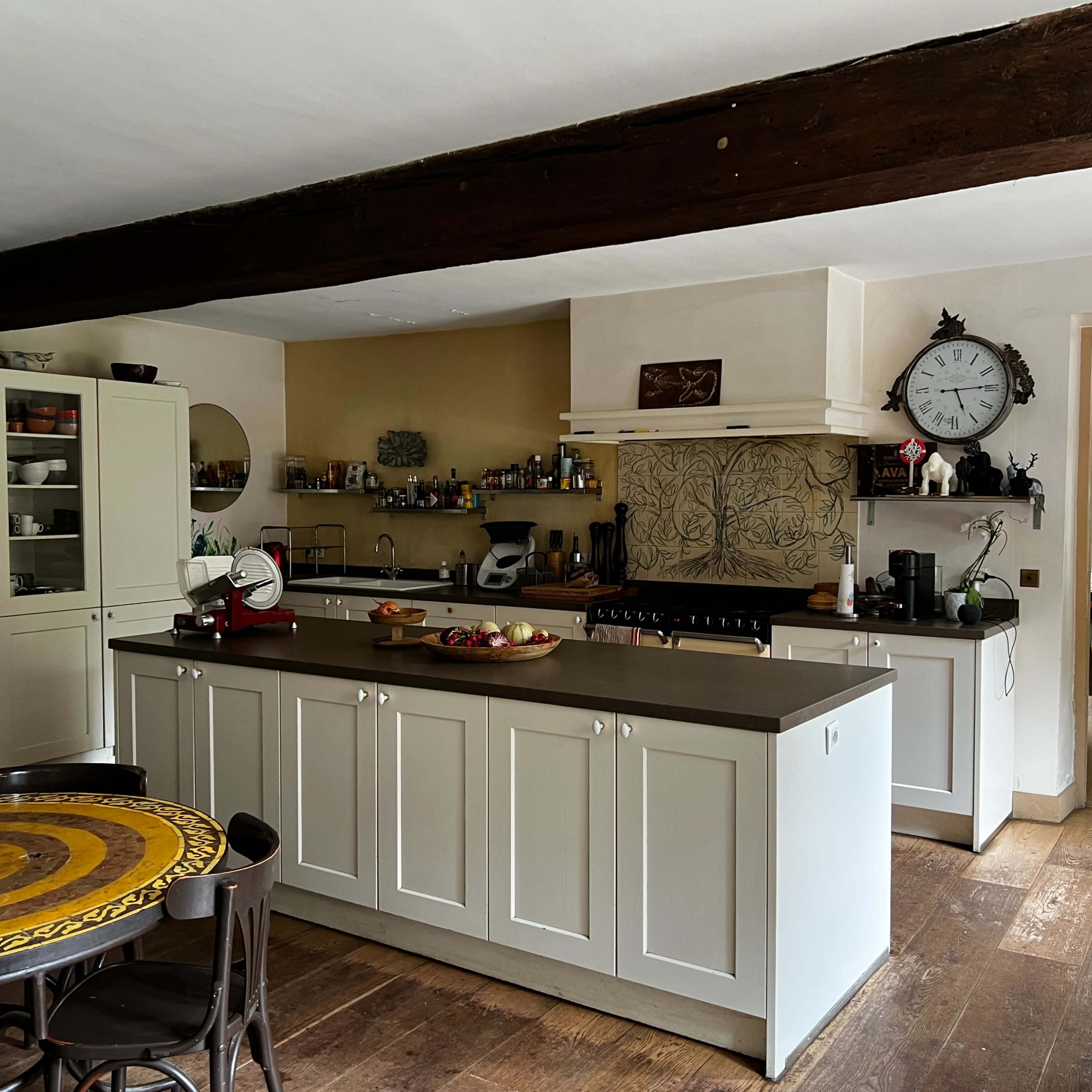 A spacious kitchen with white cabinetry, a central island, wooden beams, and a large clock on the wall.