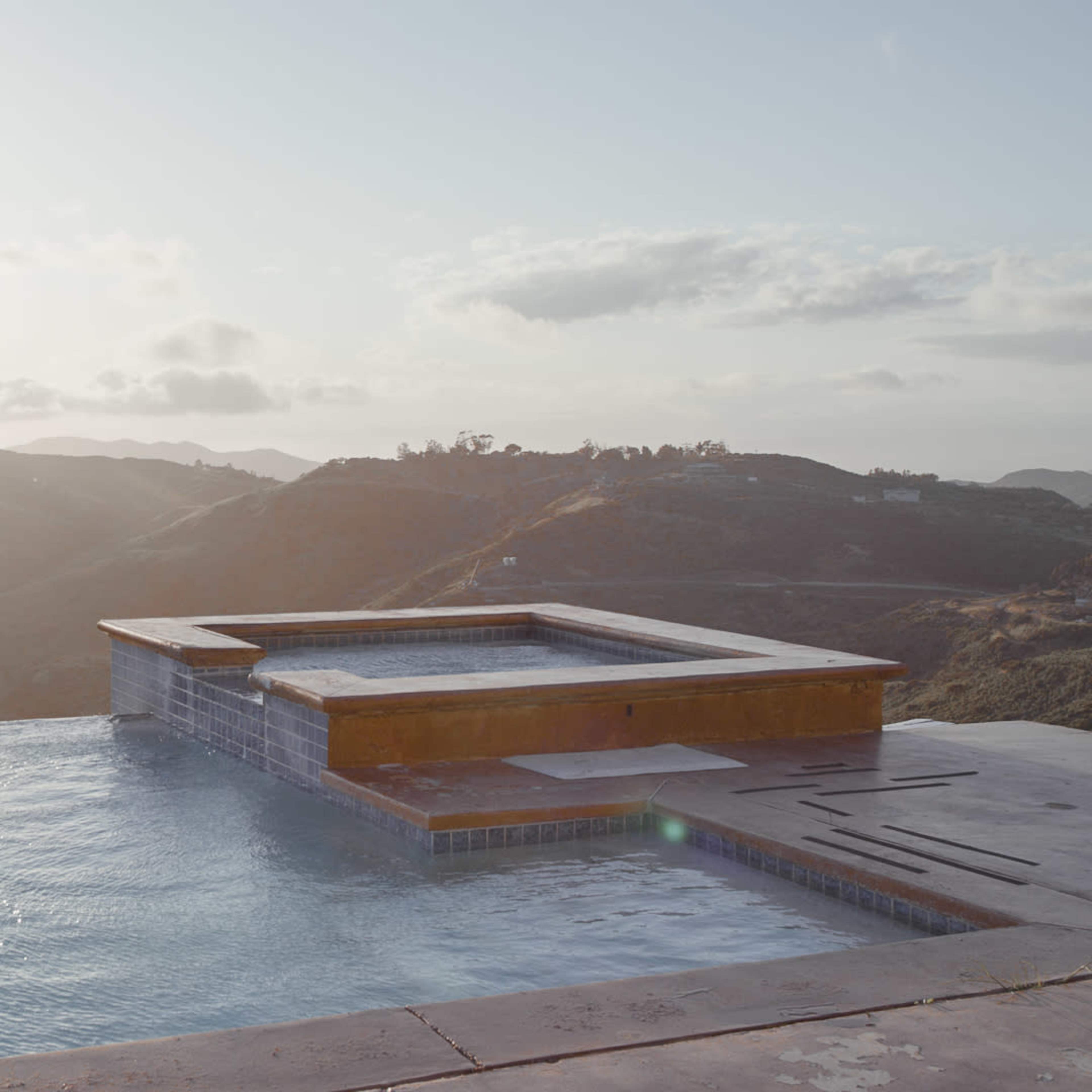 A hot tub sits at the edge of an infinity pool overlooking a mountainous landscape at sunset.