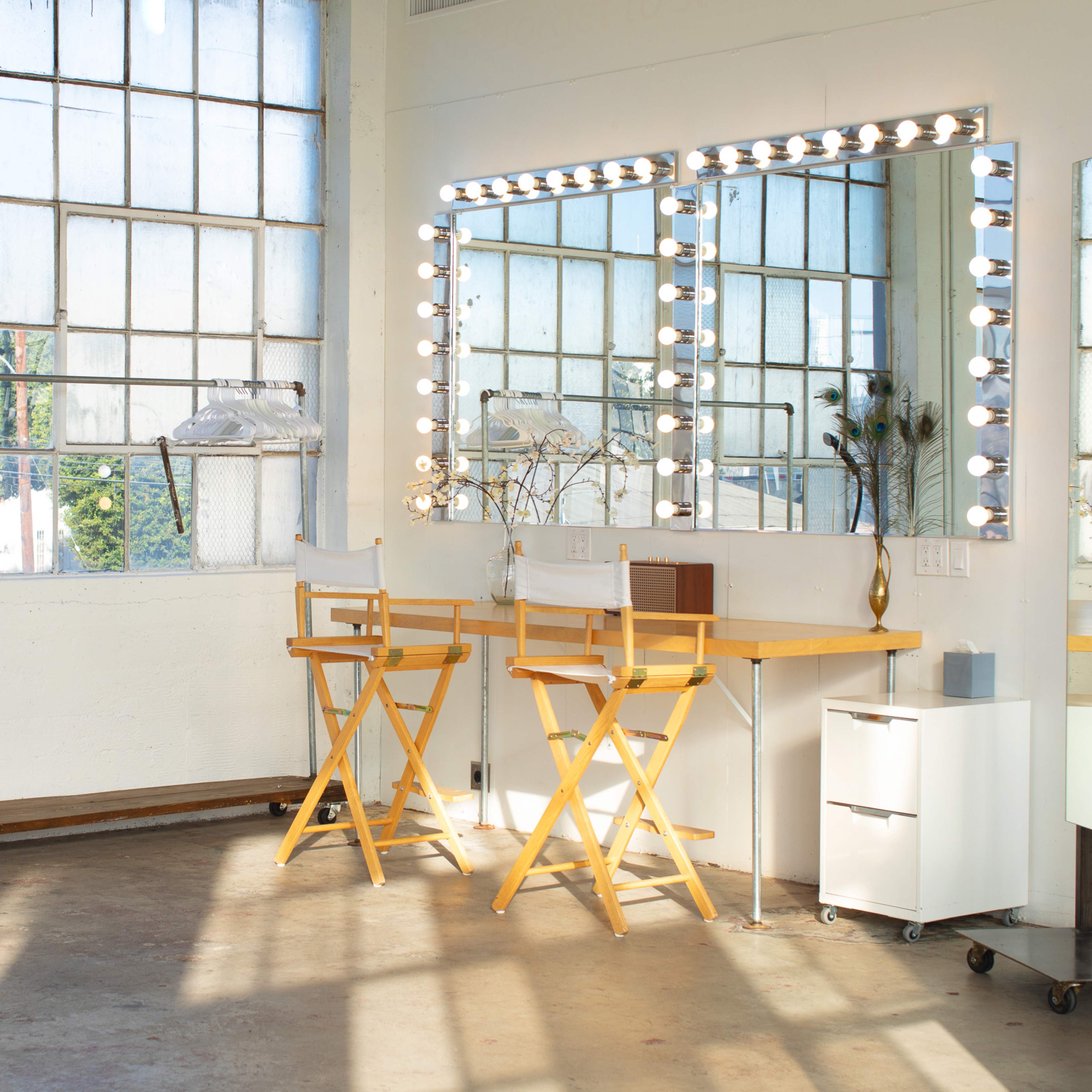 A bright, minimalist makeup room featuring two wooden director's chairs, a mirror with bulbs, and a white cabinet against a wall of large windows.