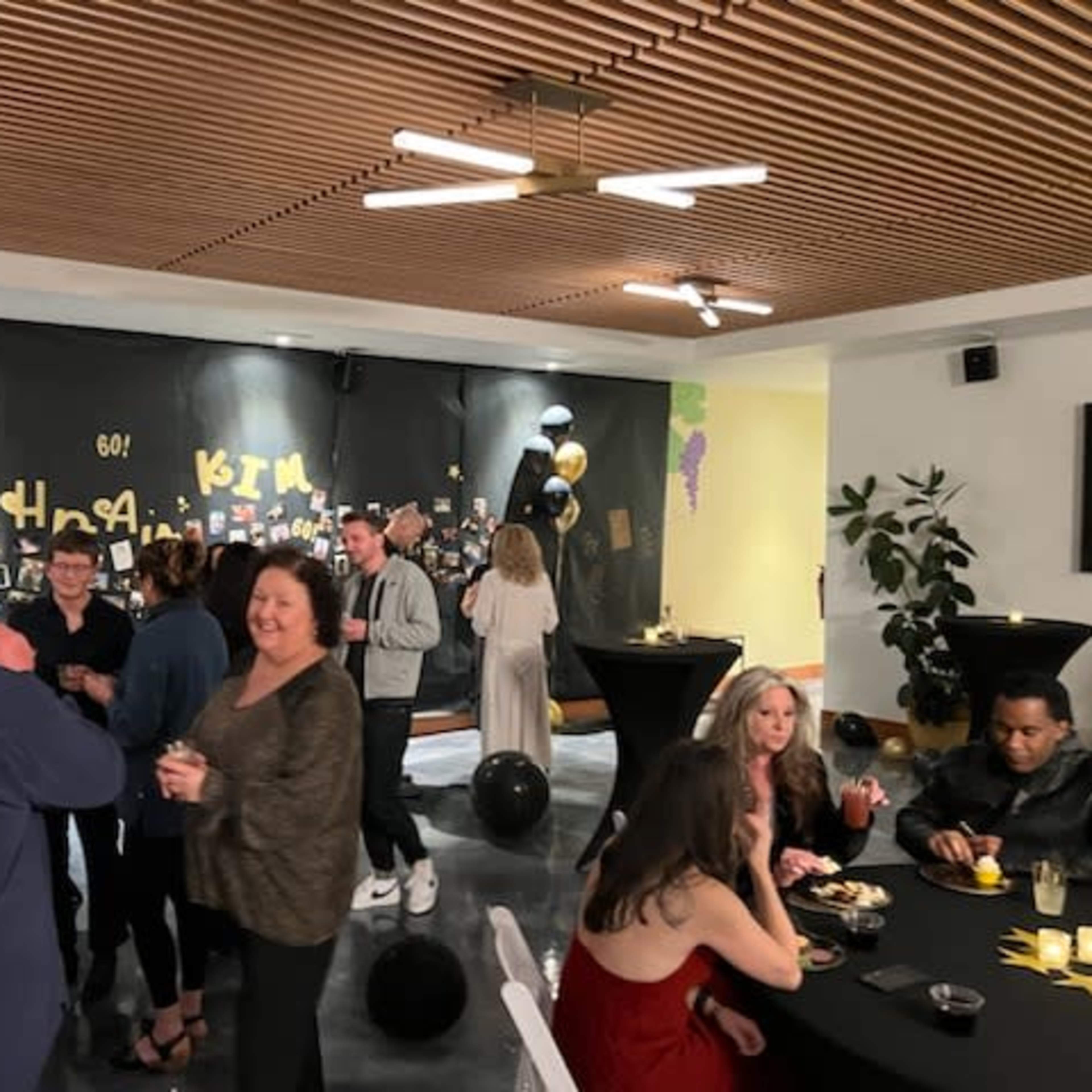 A group of people socializes at a birthday party, with decorations and a dark wall displaying the message "Happy Birthday Kim."