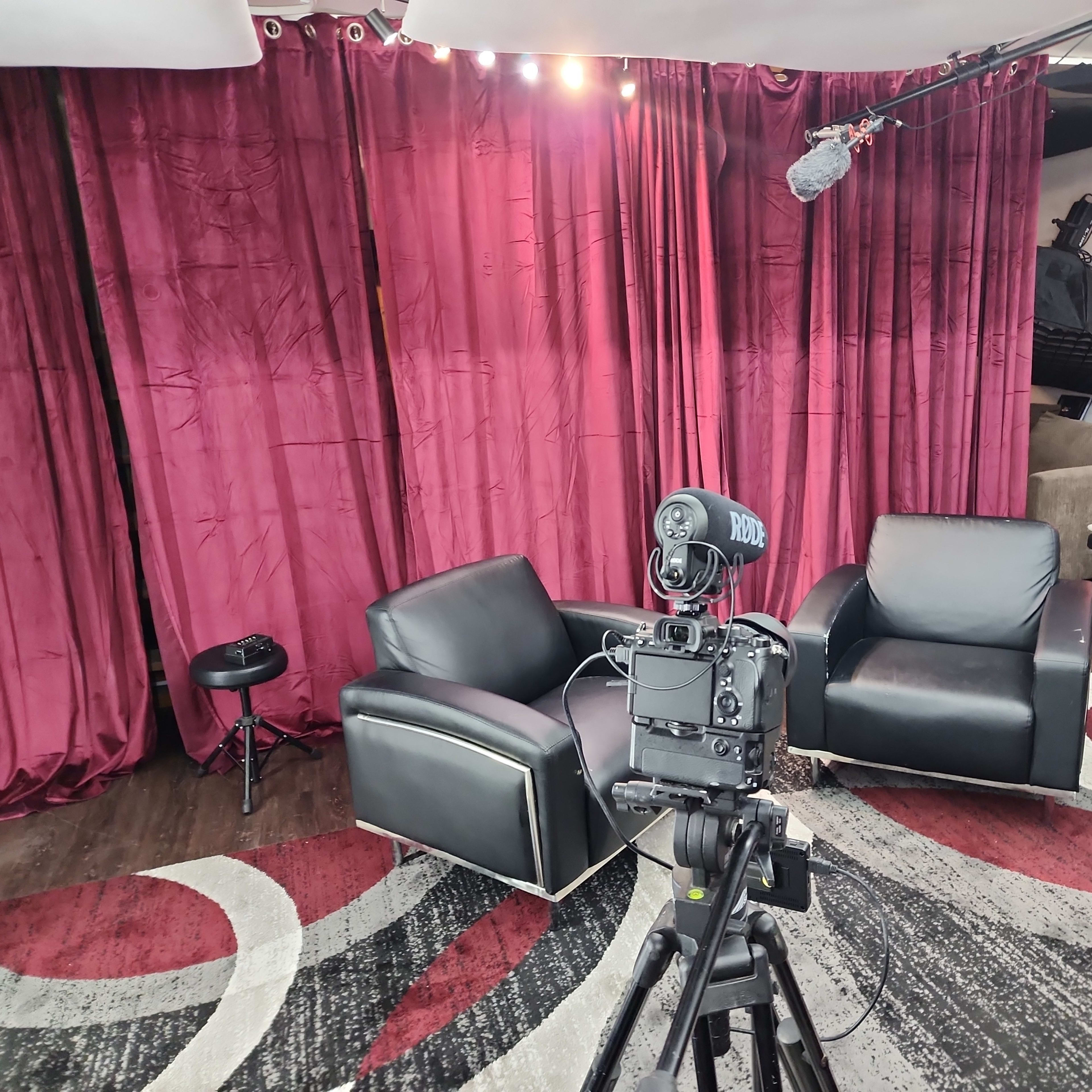 A recording setup with two black chairs in front of a red curtain, a microphone overhead, and a camera on a tripod.