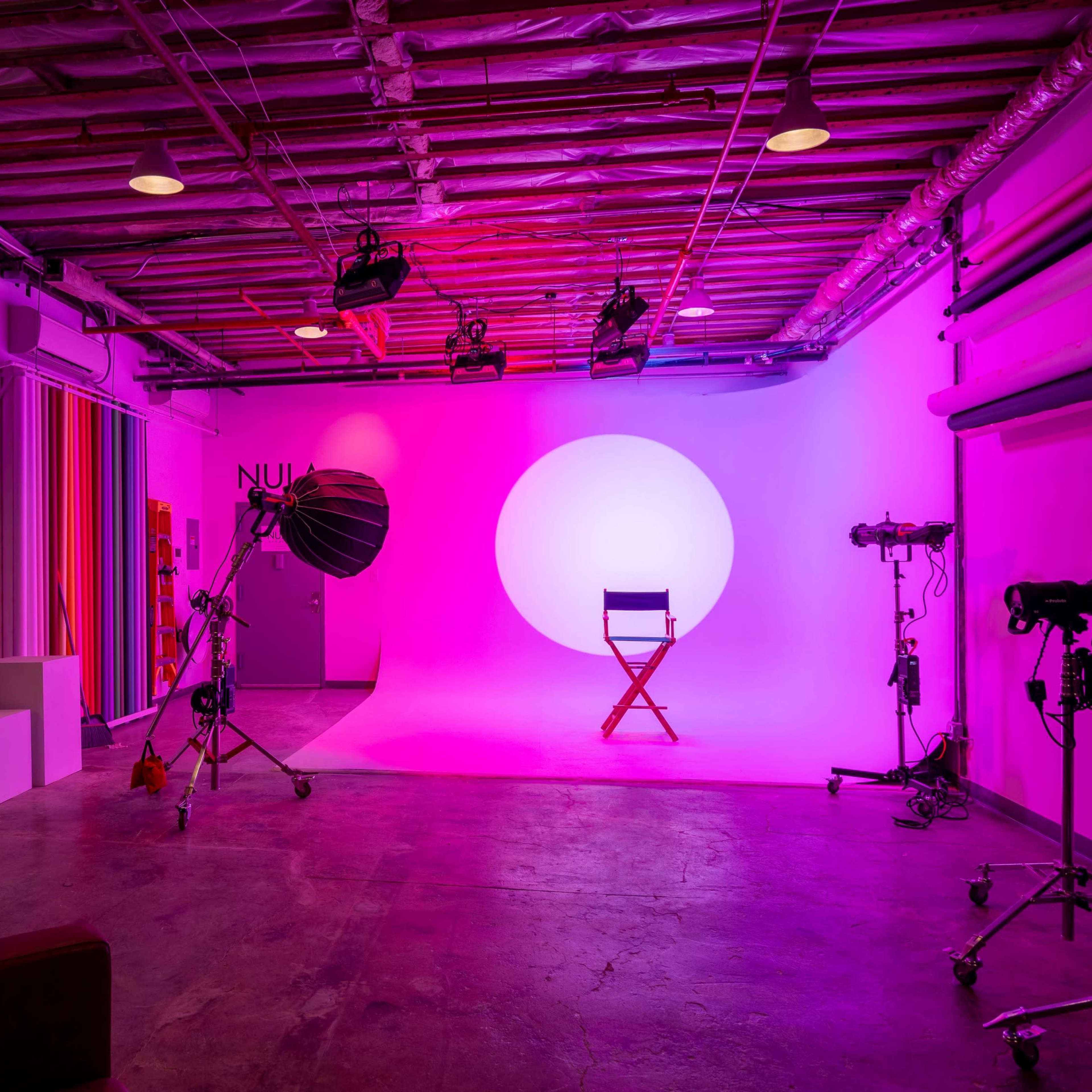 A photography studio features a brightly lit backdrop with a circular light, various lighting equipment, and a director's chair in the center.