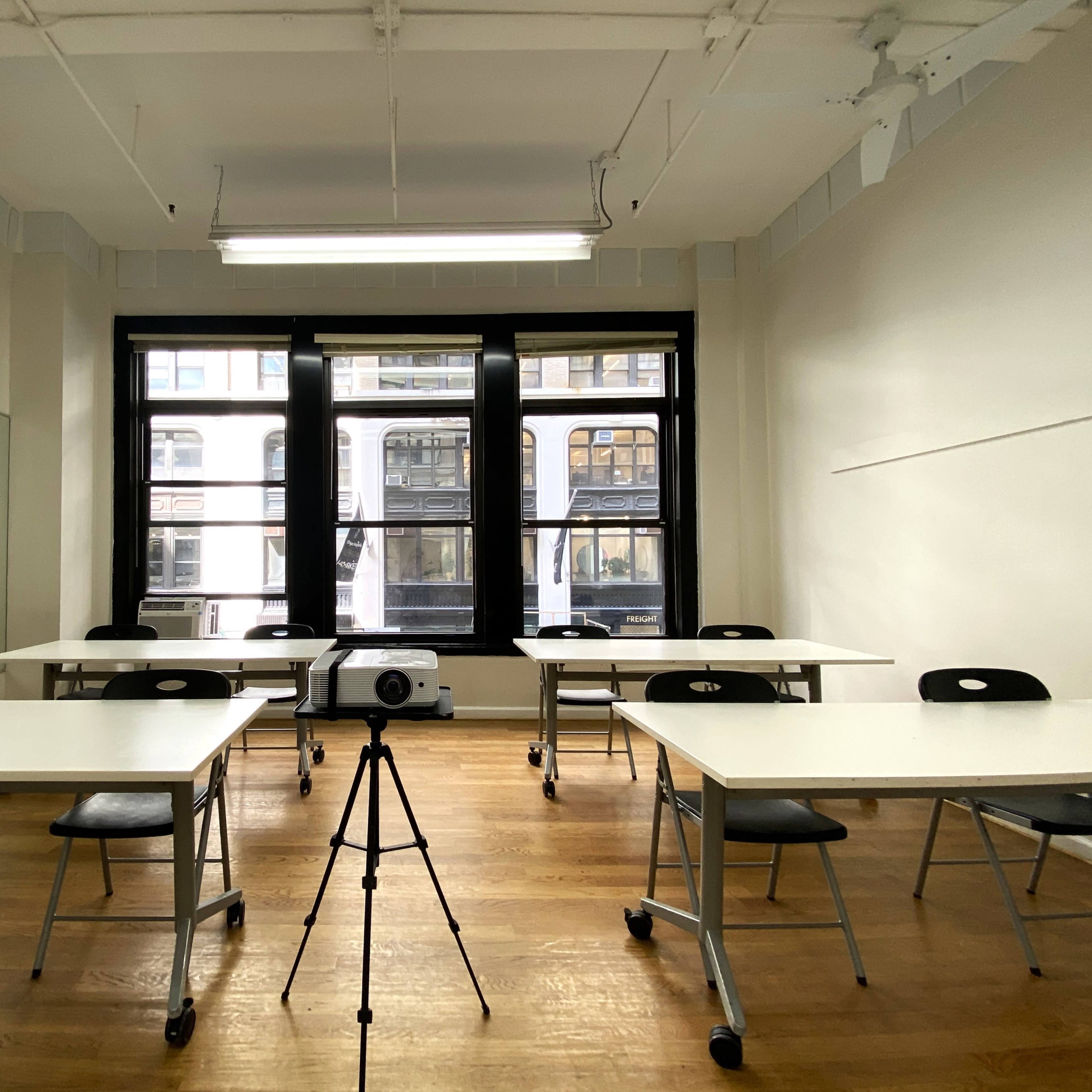 A bright, empty classroom is set up with several tables and chairs facing a projector on a tripod, with large windows providing a view of the street outside.