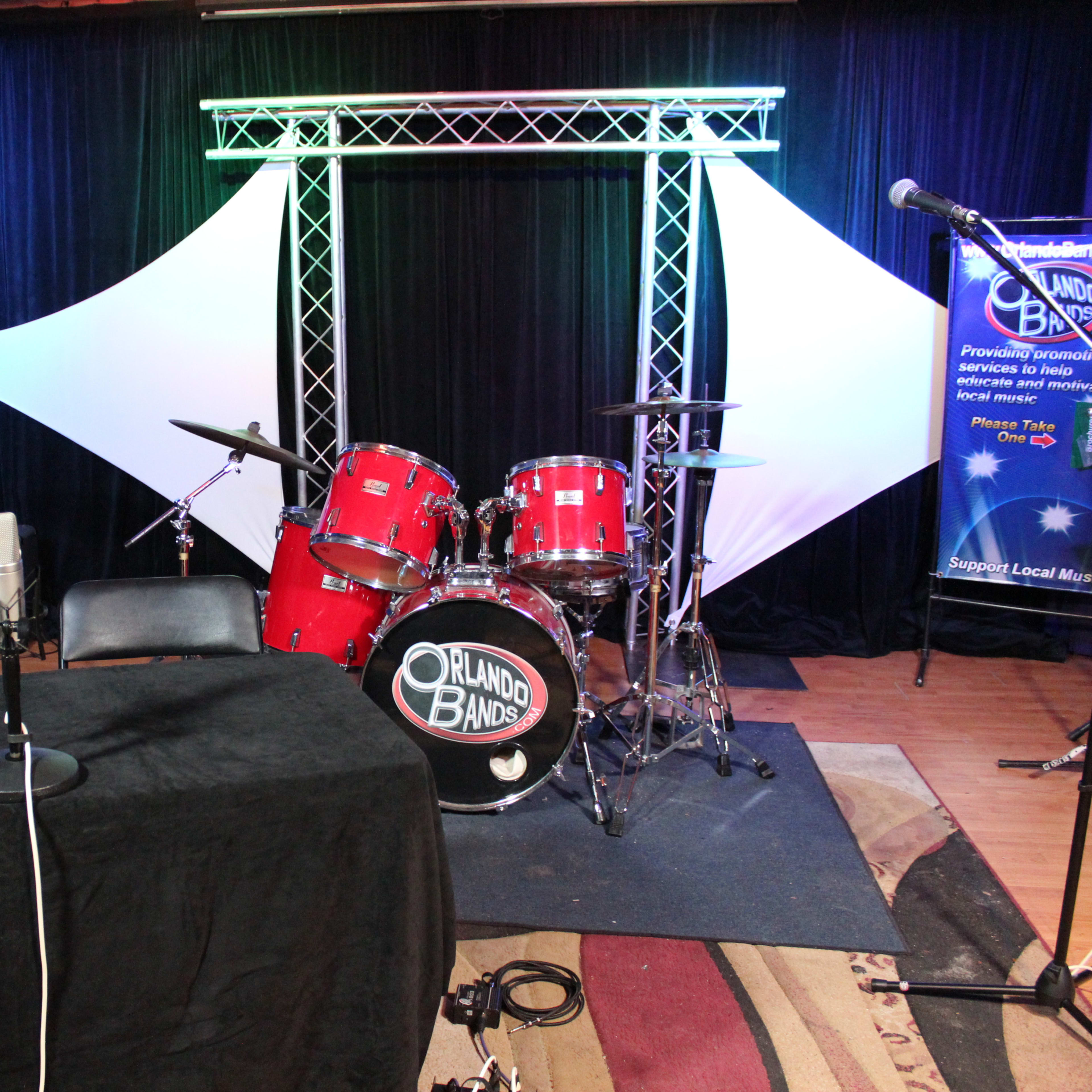 A small stage setup with a drum kit, microphones, and a laptop on a table, surrounded by black curtains and lighting.