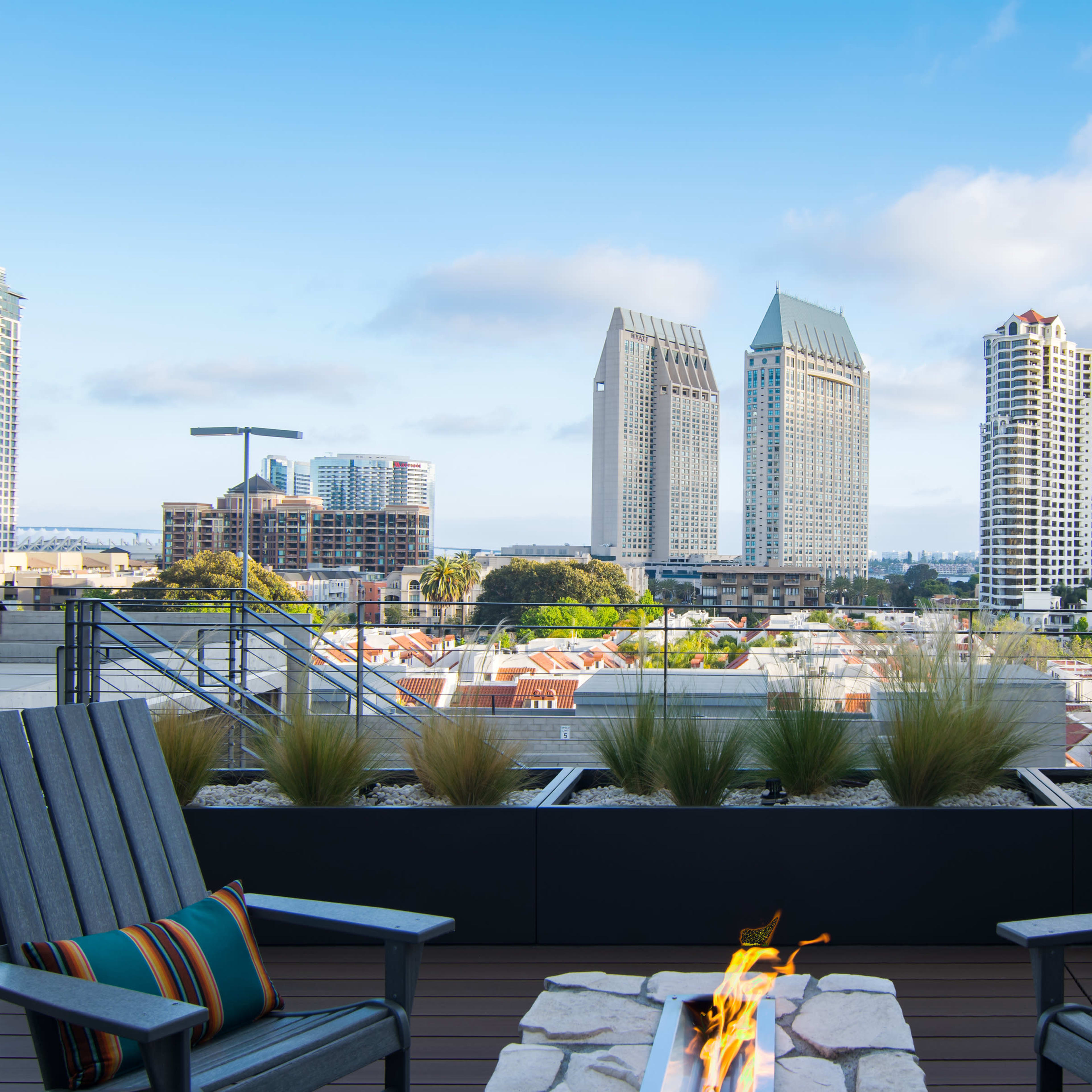 A rooftop terrace features two wooden chairs facing a fire pit, with a skyline of high-rise buildings in the background.