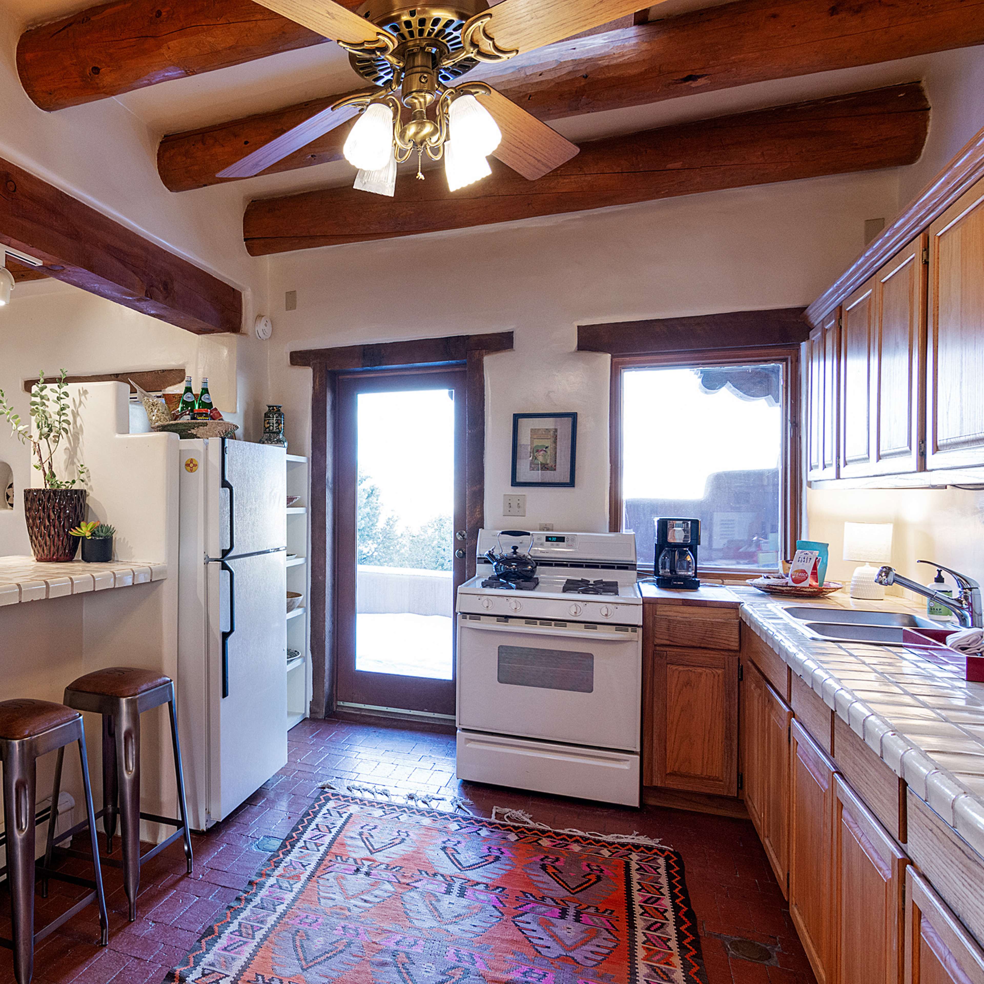 A rustic kitchen features wooden beams, a white tile countertop with bar stools, and a view of a patio through the doorway.