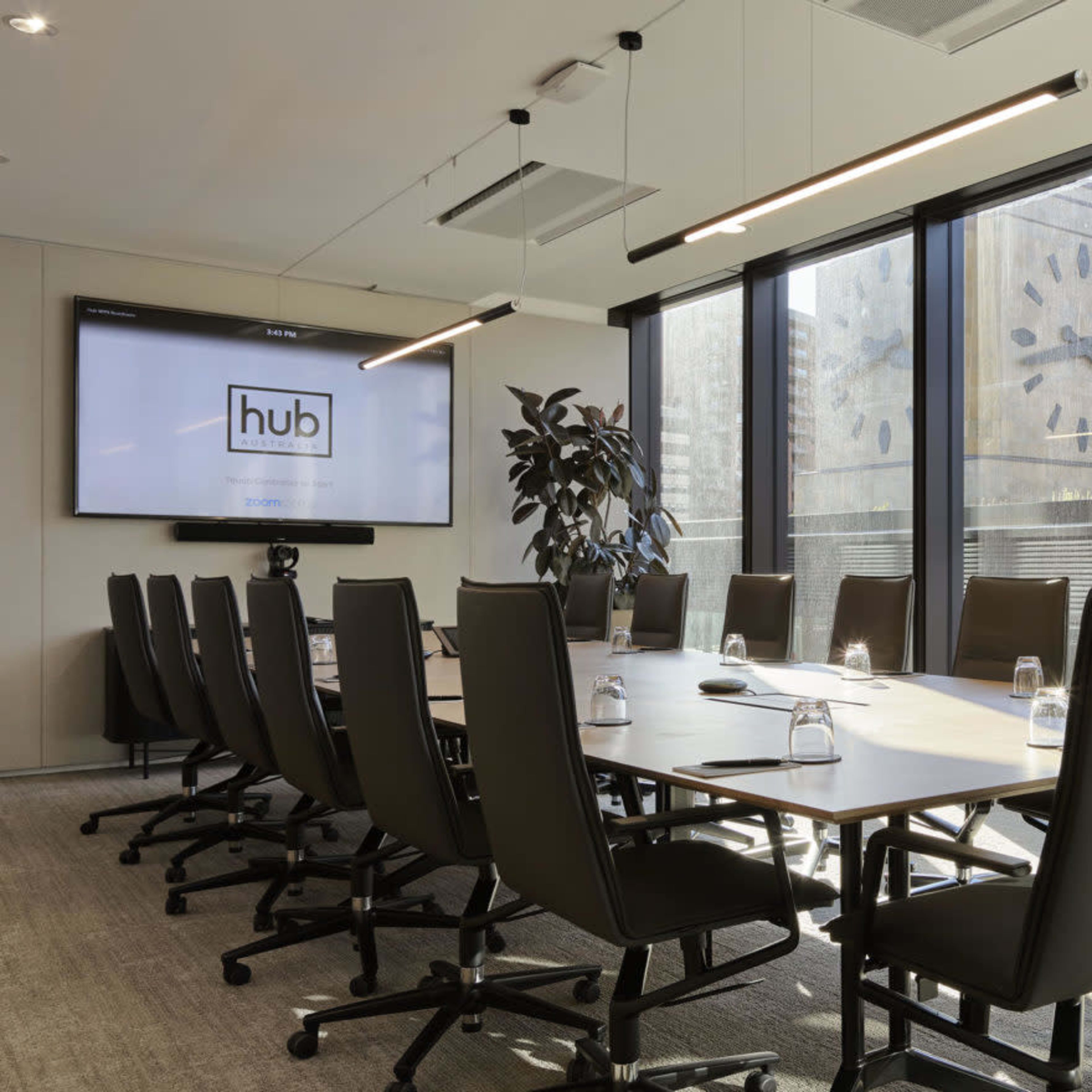 A modern conference room features a long table with black ergonomic chairs, large windows revealing a cityscape, and a wall-mounted screen displaying the logo of "hub."