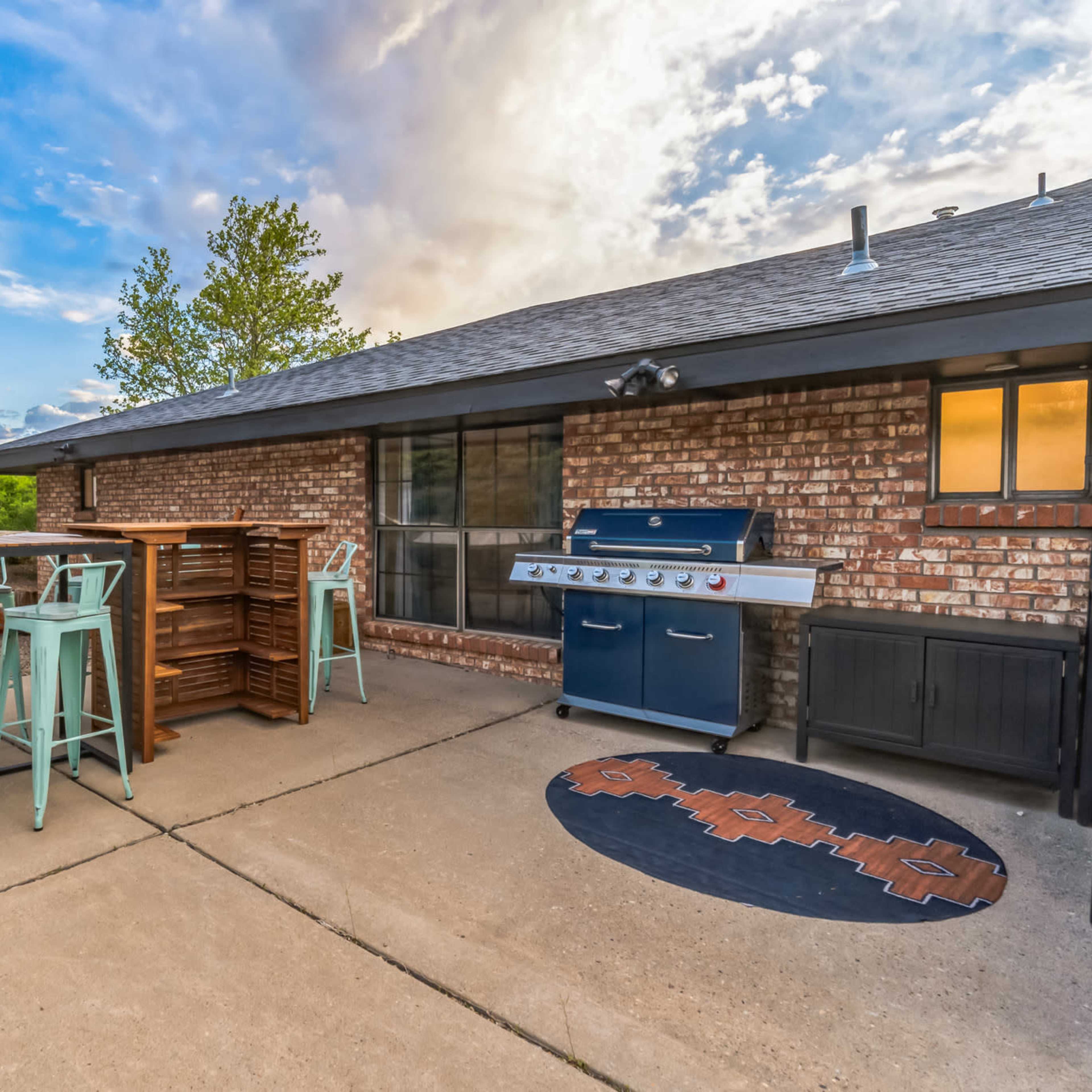 A patio area featuring a blue grill, high-top tables with stools, and a backdrop of trees and clouds.