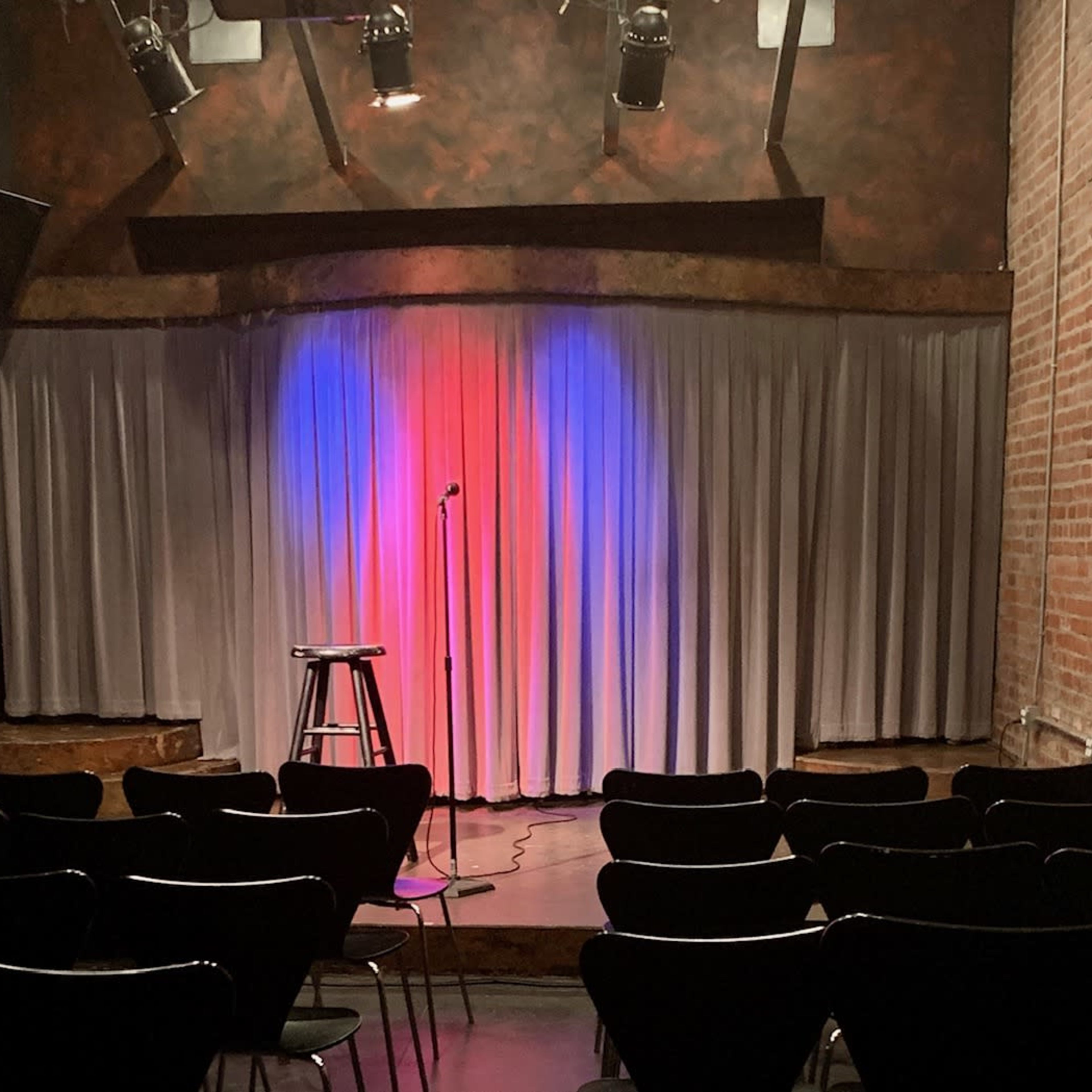 A small comedy stage with a microphone stand and a tall stool in front of a curtain illuminated with red and blue lights.