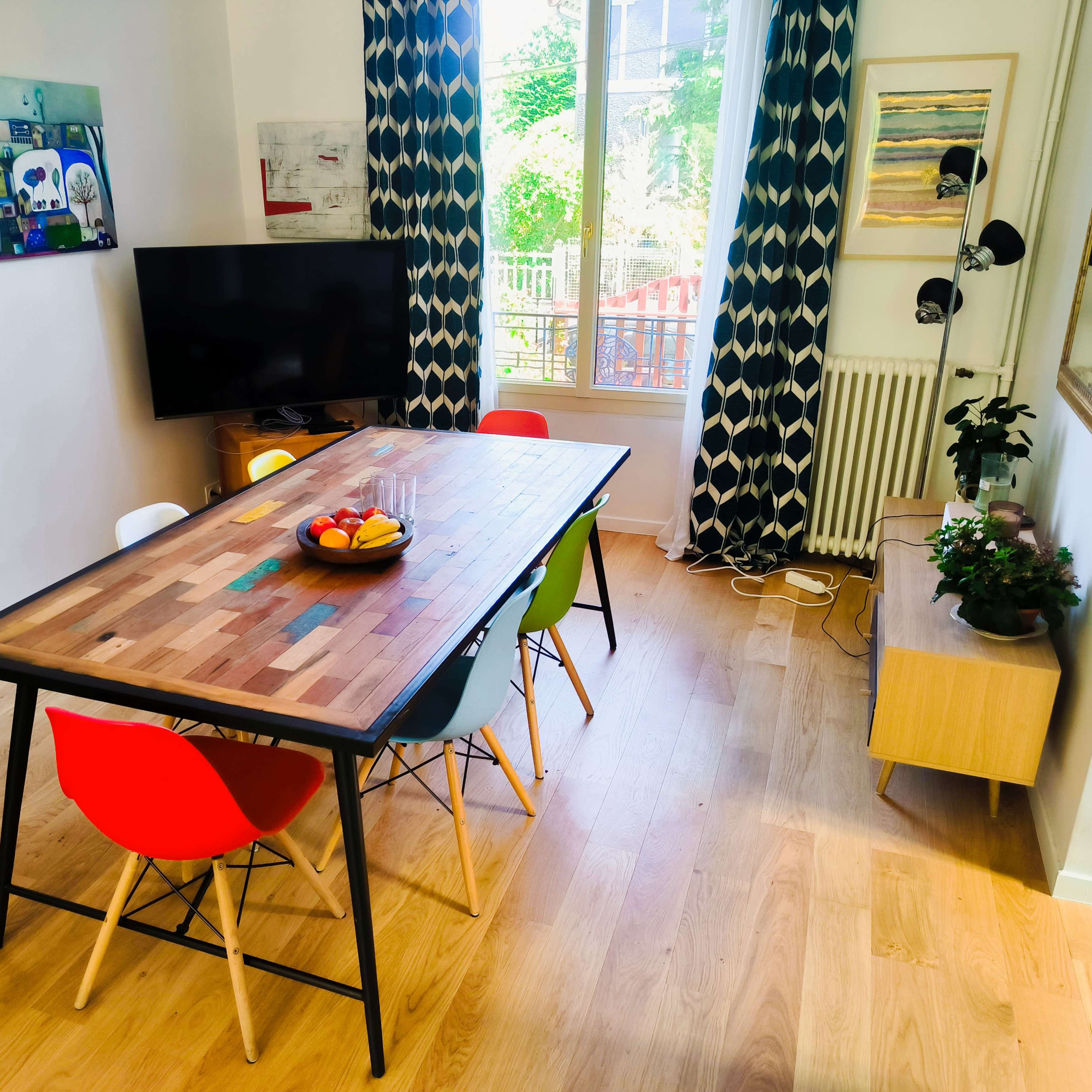 A dining area features a wooden table surrounded by colorful chairs, a television on the wall, and large windows allowing natural light to enter.