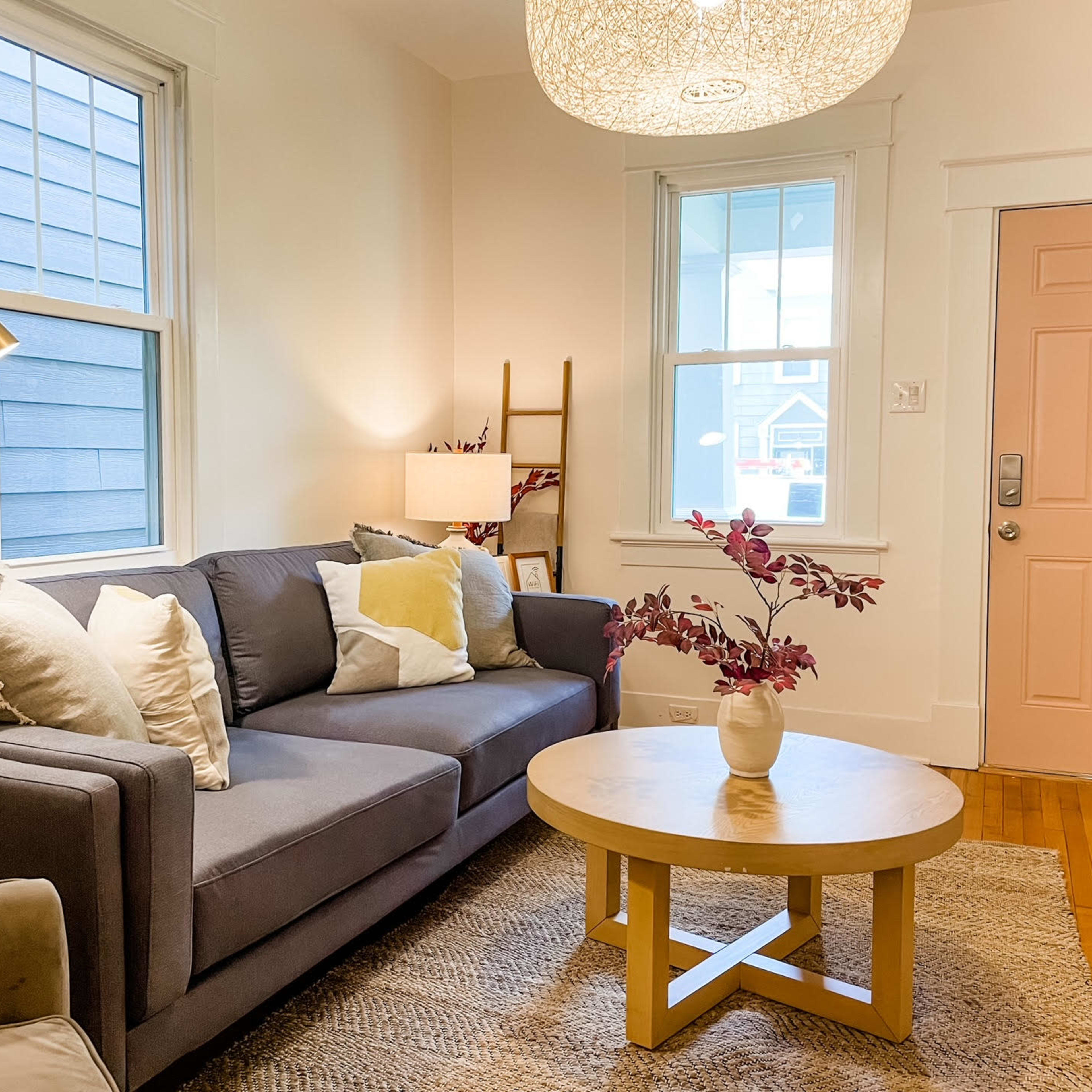 A gray sofa with decorative pillows faces a round coffee table and a light fixture in a brightly lit living room with large windows and a pink door.