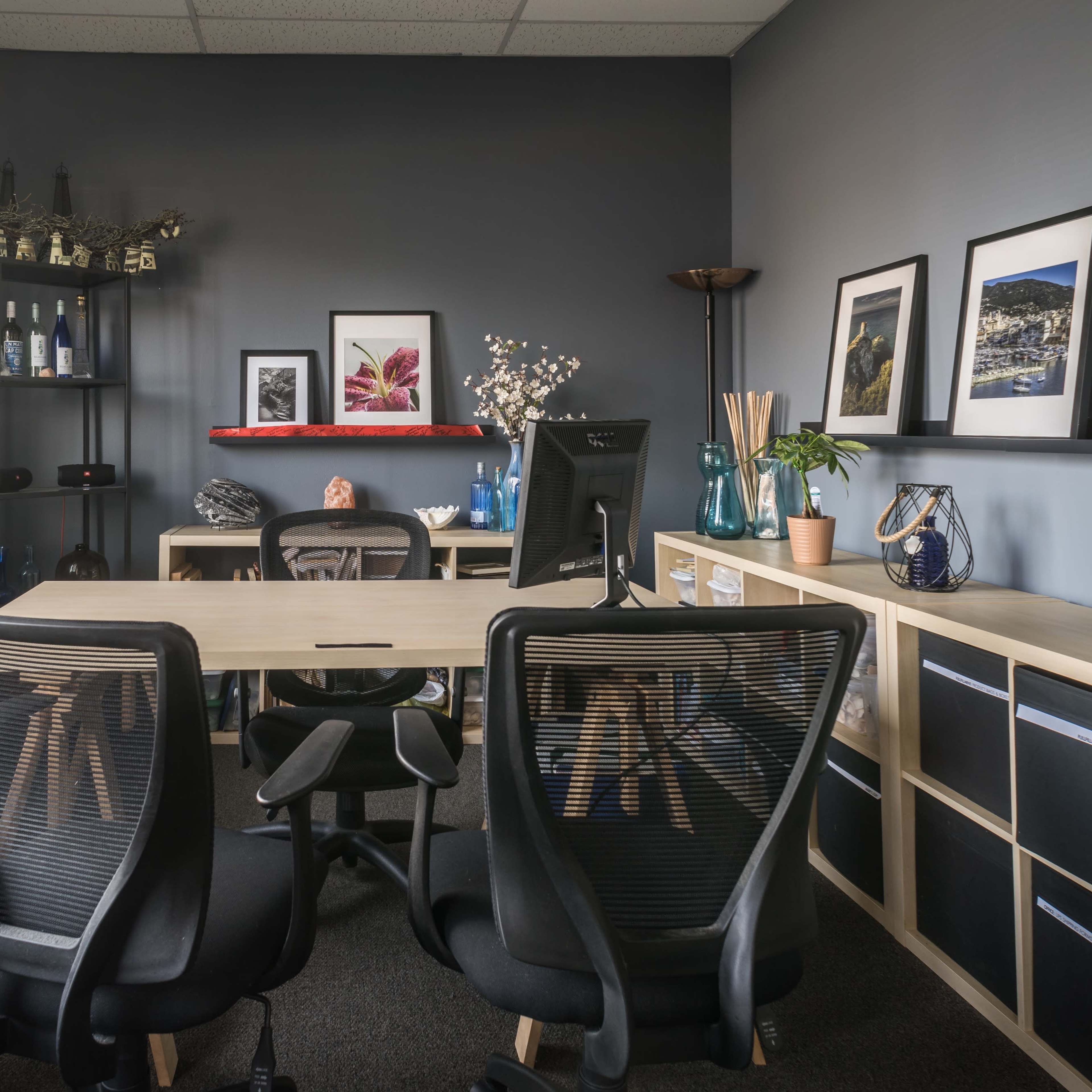 A modern office space features a desk with black chairs, shelves displaying decorative items, and framed artwork on the walls.