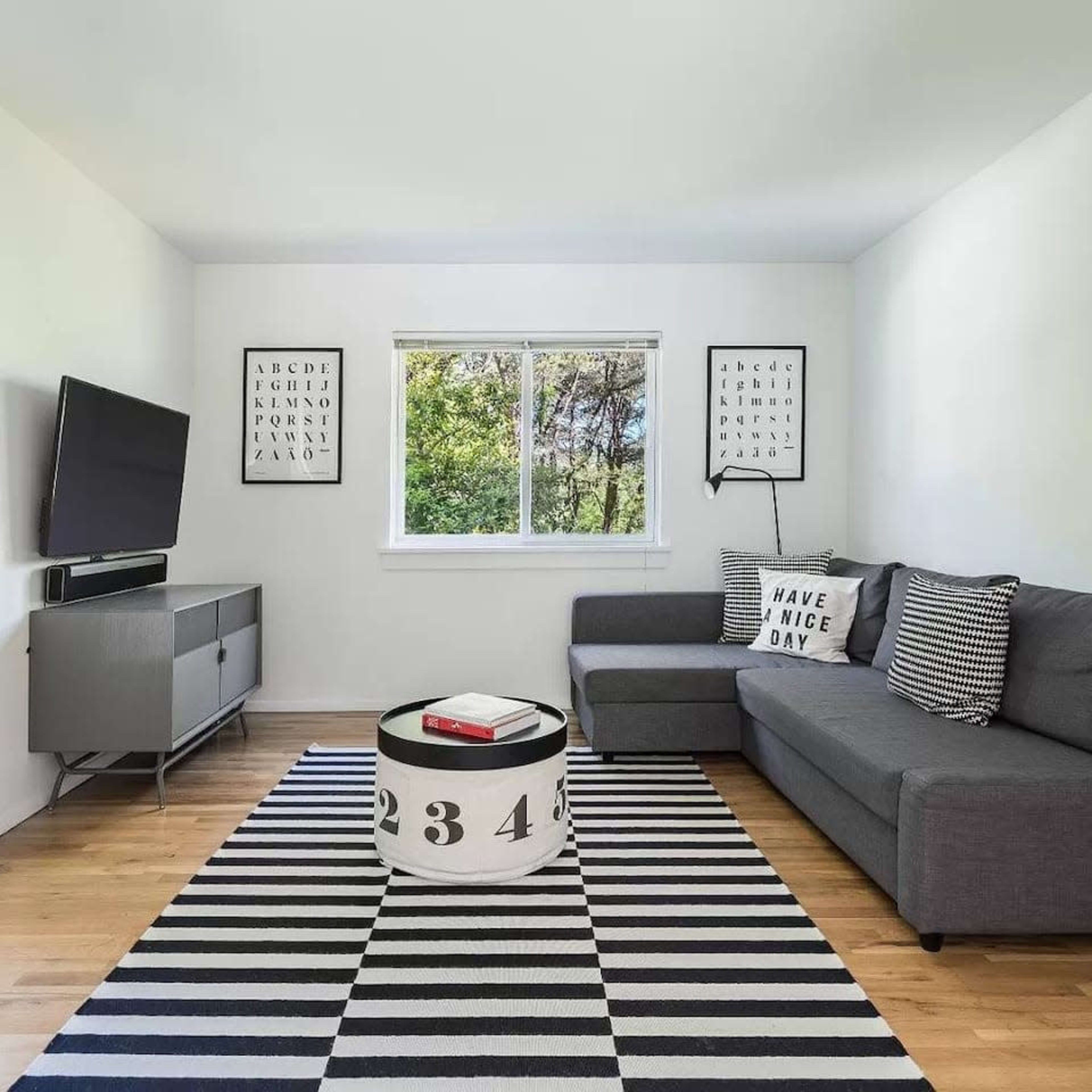 A modern living room featuring a gray sectional sofa, a black and white striped rug, a coffee table with bold numbers, and a television mounted on the wall.