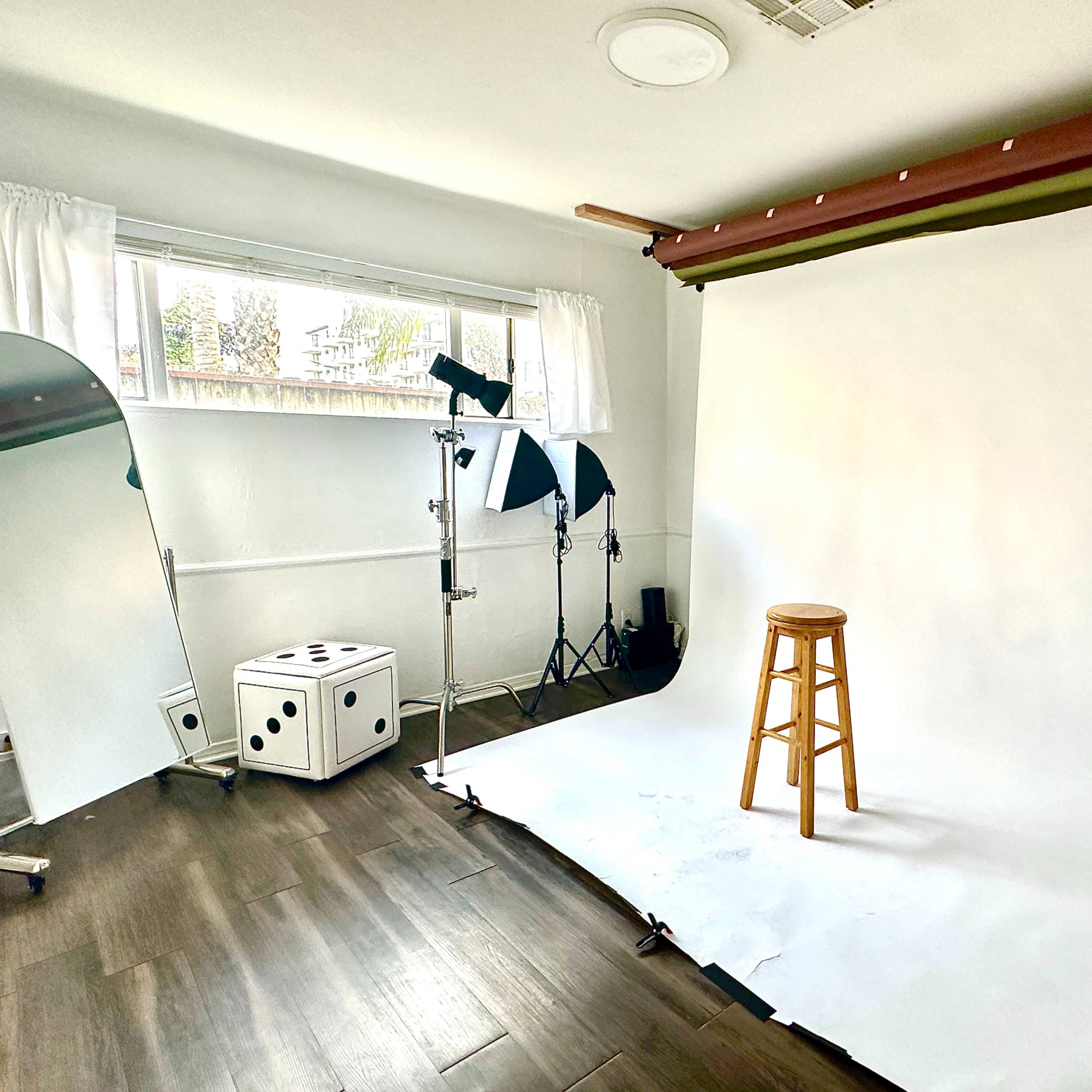 A photography studio with a blank backdrop, a wooden stool, lighting equipment, and a mirror positioned to the side.