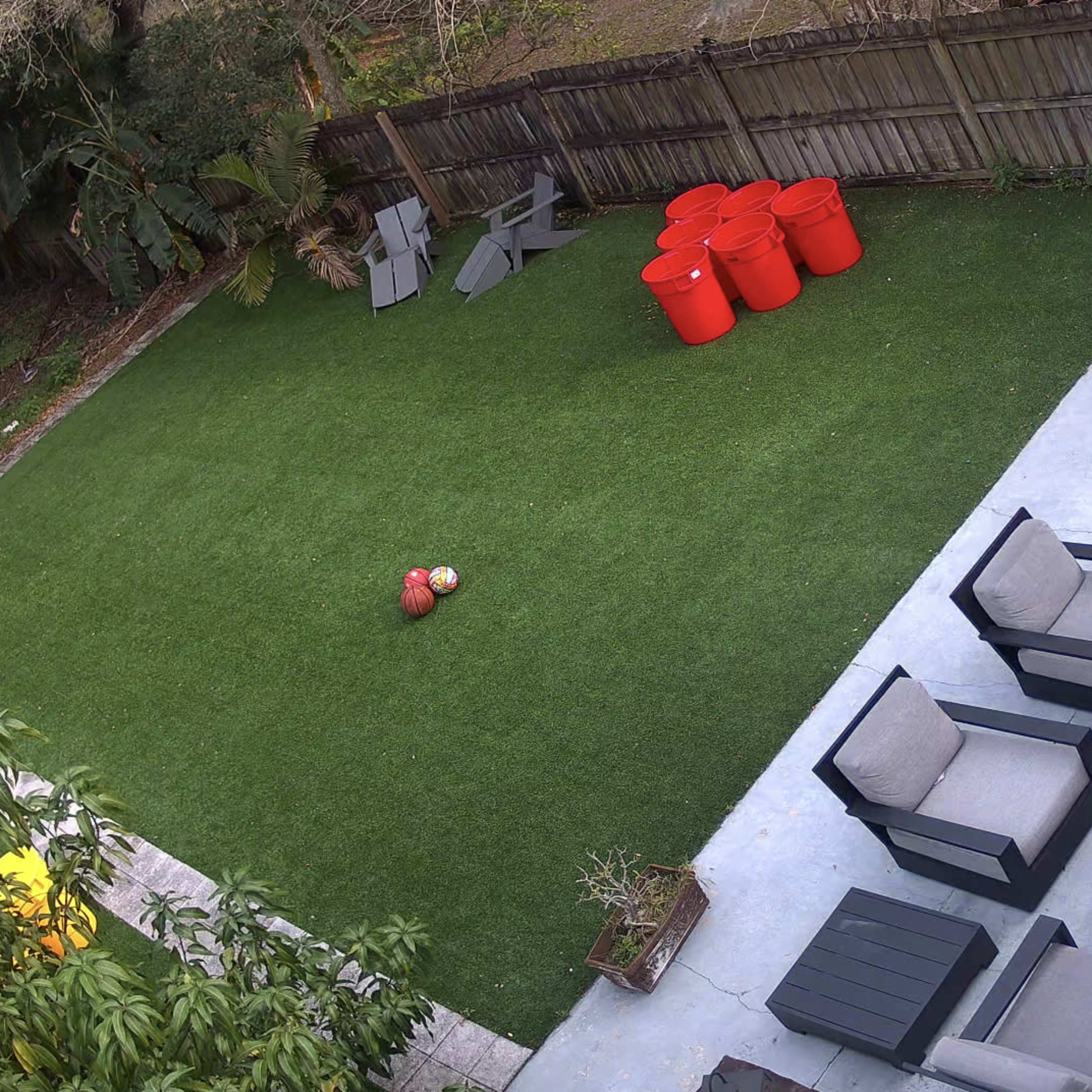 A backyard with a green artificial lawn, a seating area with gray patio furniture, and several orange barrels arranged on one side.