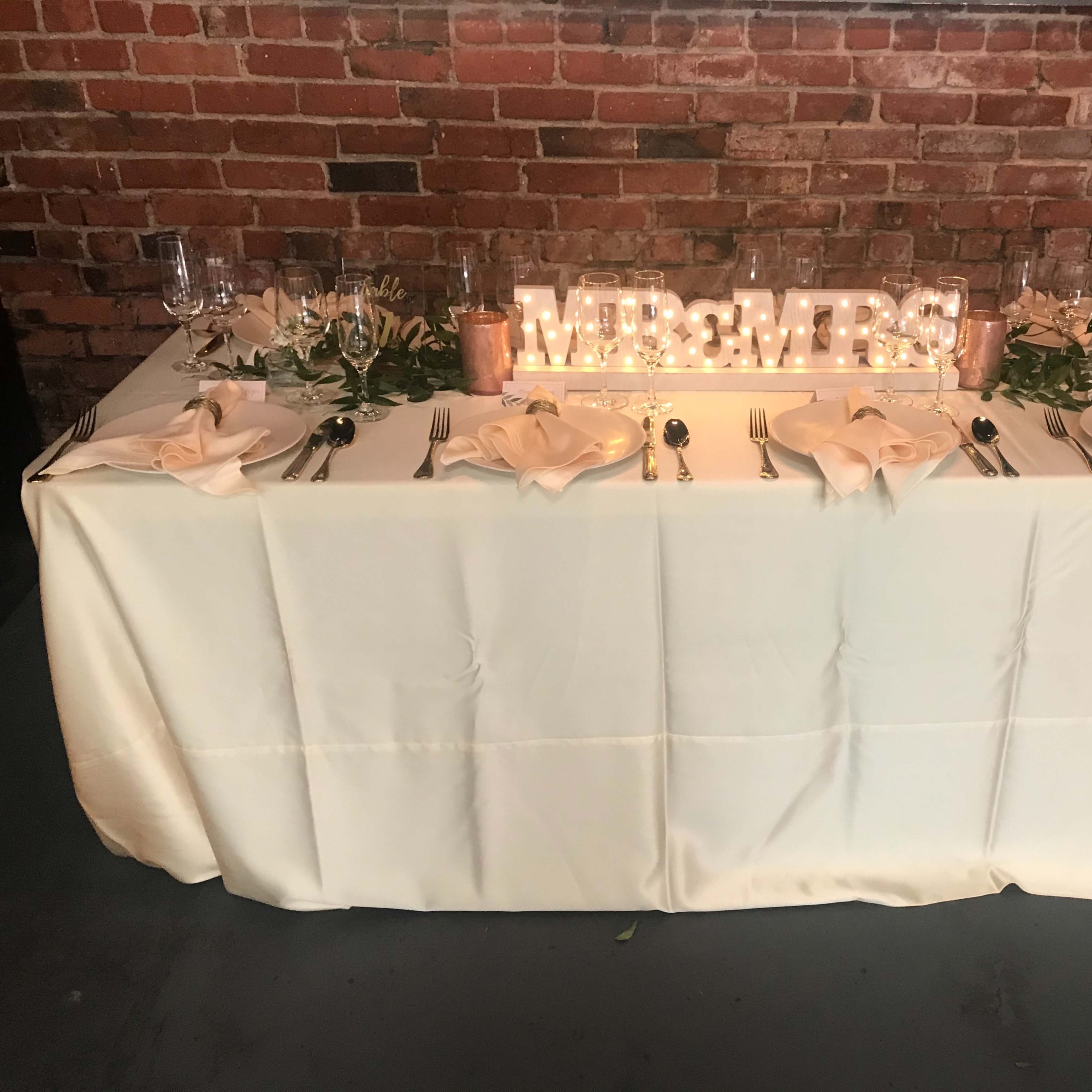 A elegantly set dining table with a light-colored tablecloth, adorned with a floral centerpiece, candles, and a decorative sign that reads "MR & MRS."