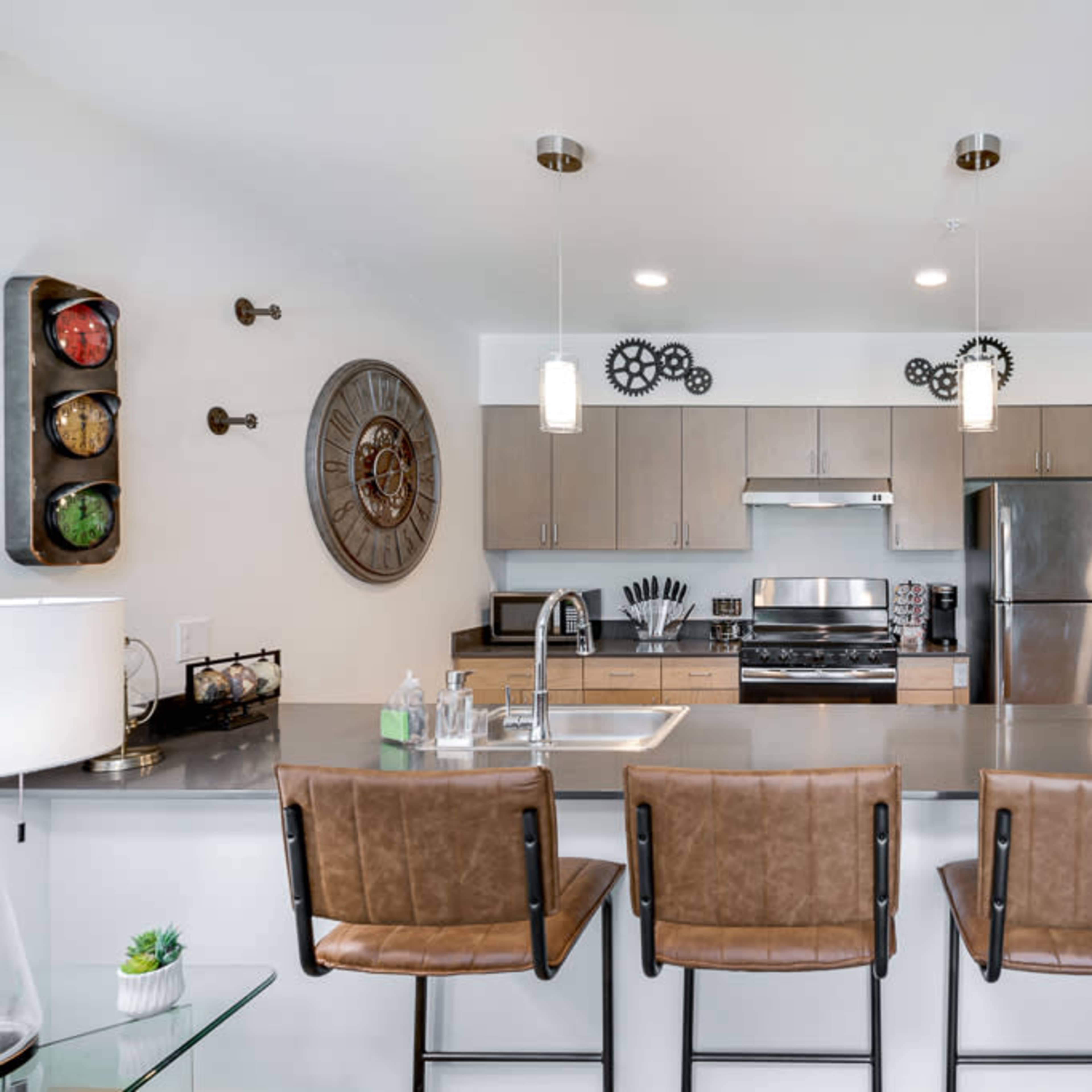 A modern kitchen features stainless steel appliances, wooden cabinets, and four brown bar stools at a gray countertop.