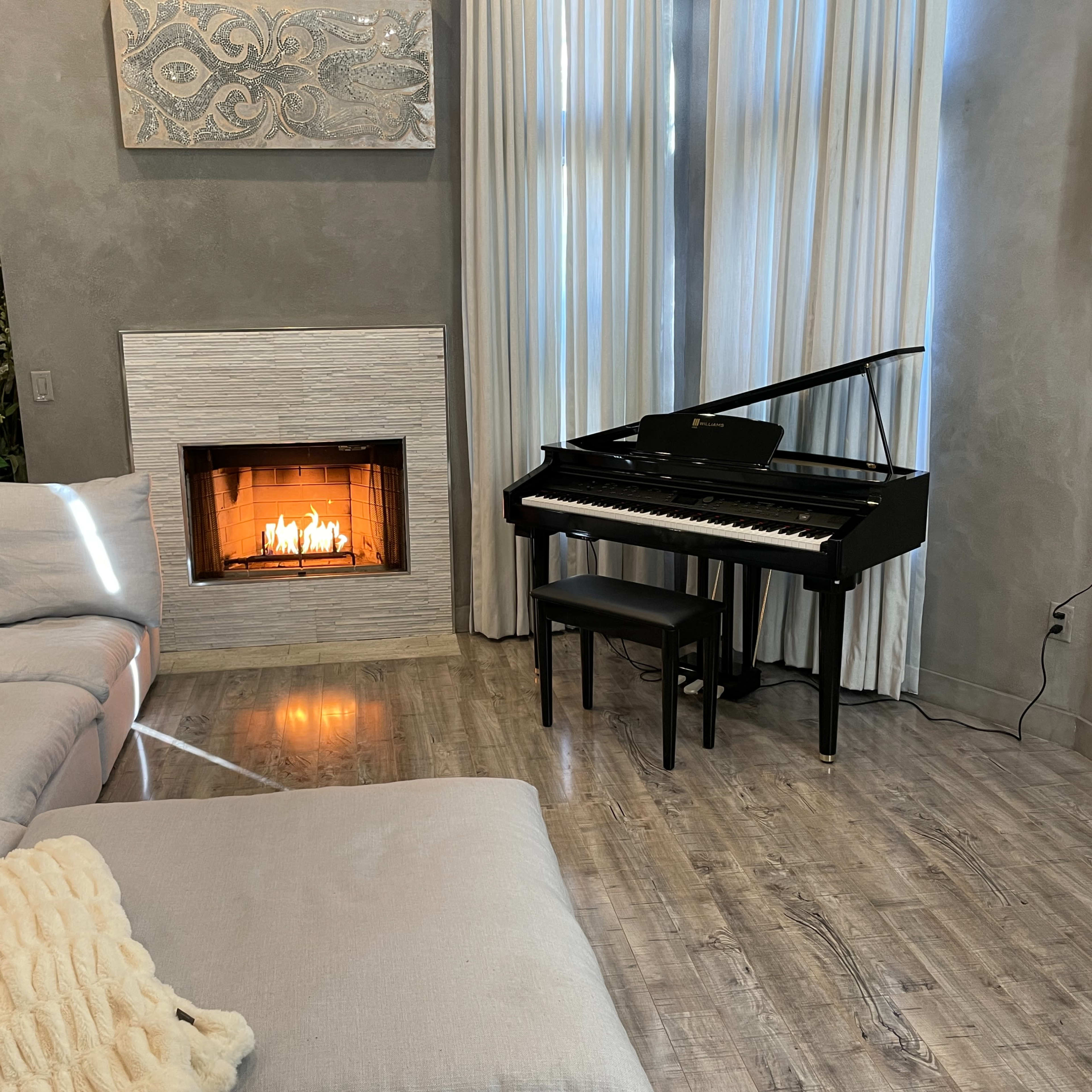 The living room features a grand piano next to a fireplace, with a sectional sofa and large windows dressed in white curtains.