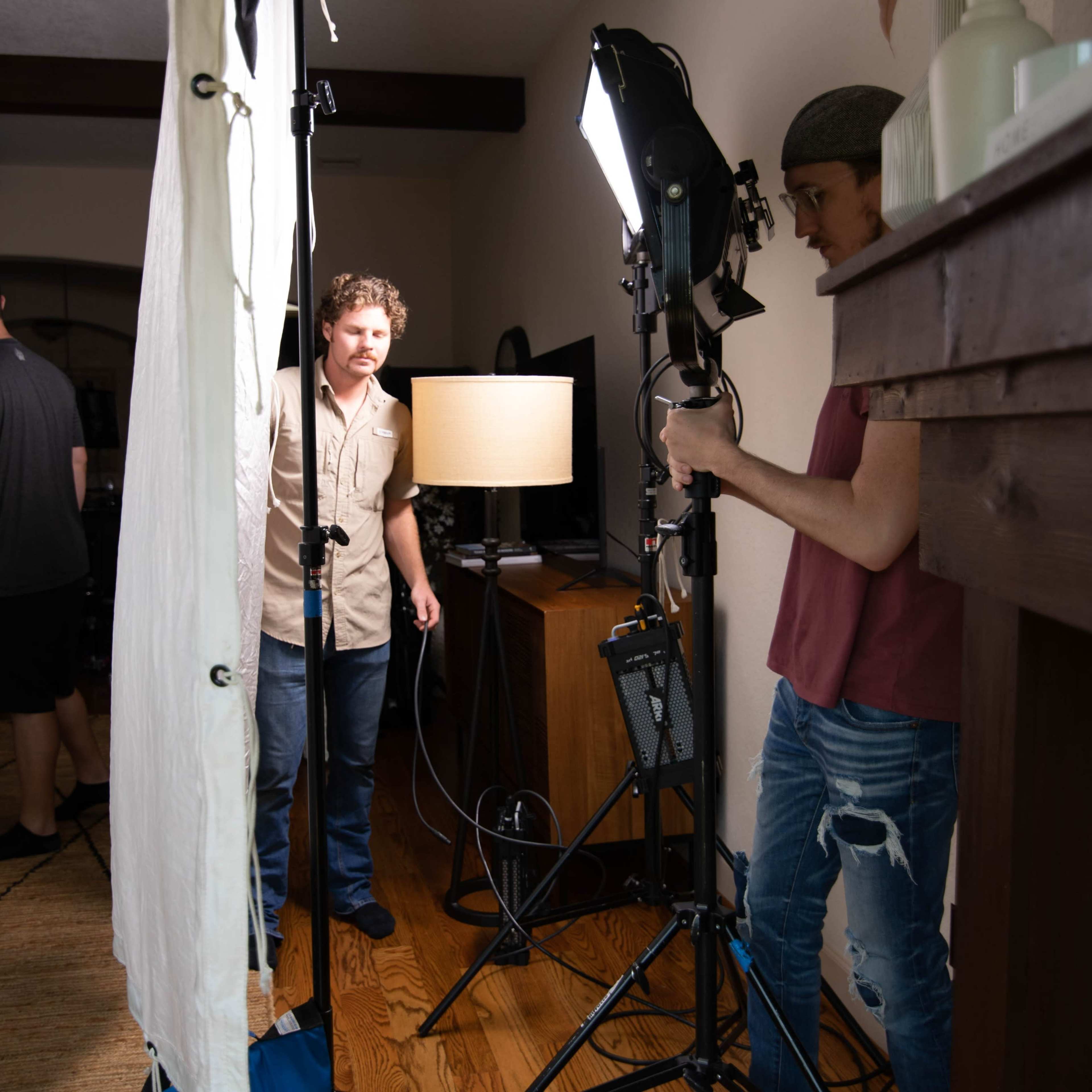 A film crew sets up lighting equipment in a living room while a few people are positioned in the background.
