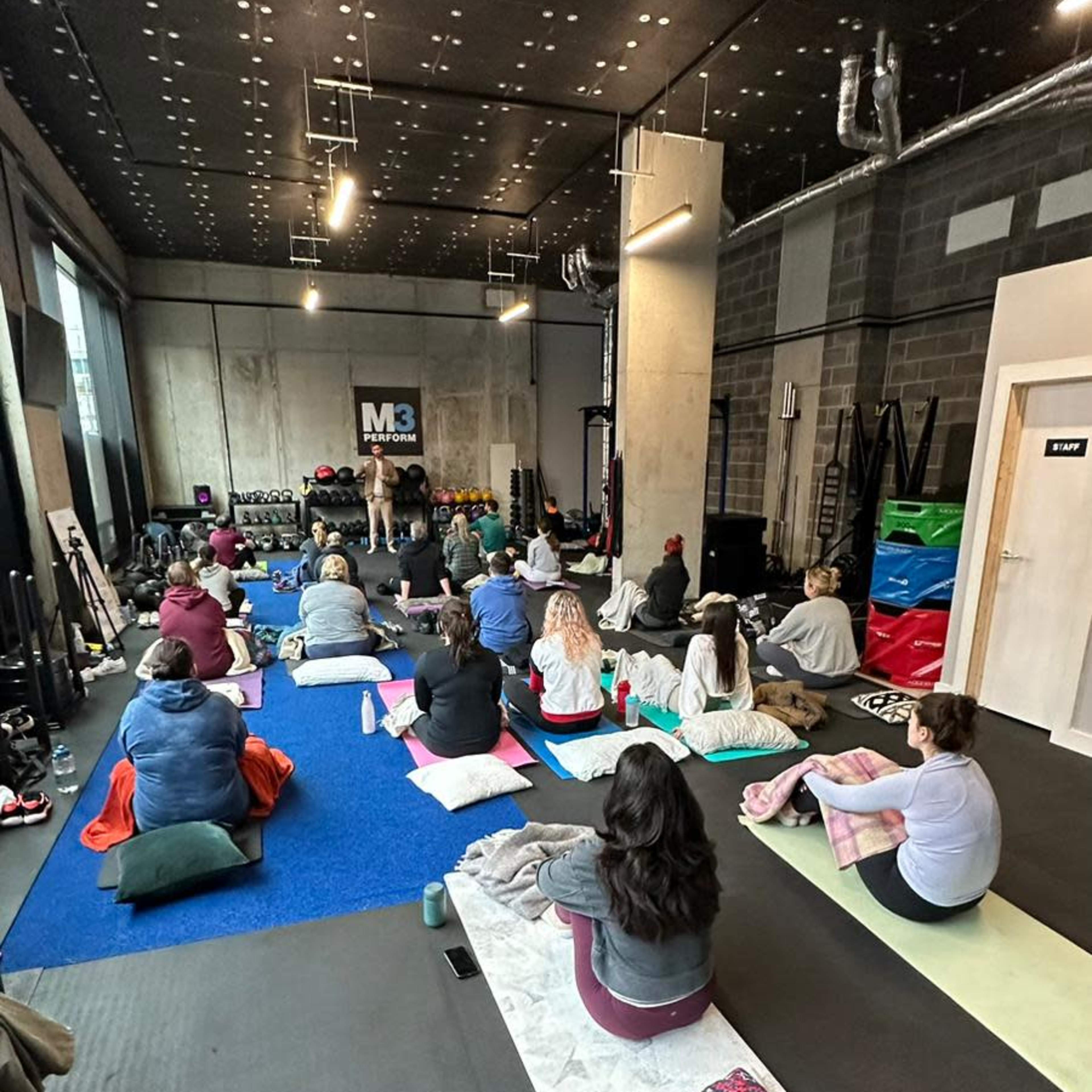 A group of individuals is seated on mats in a fitness studio, facing an instructor at the front of the room.