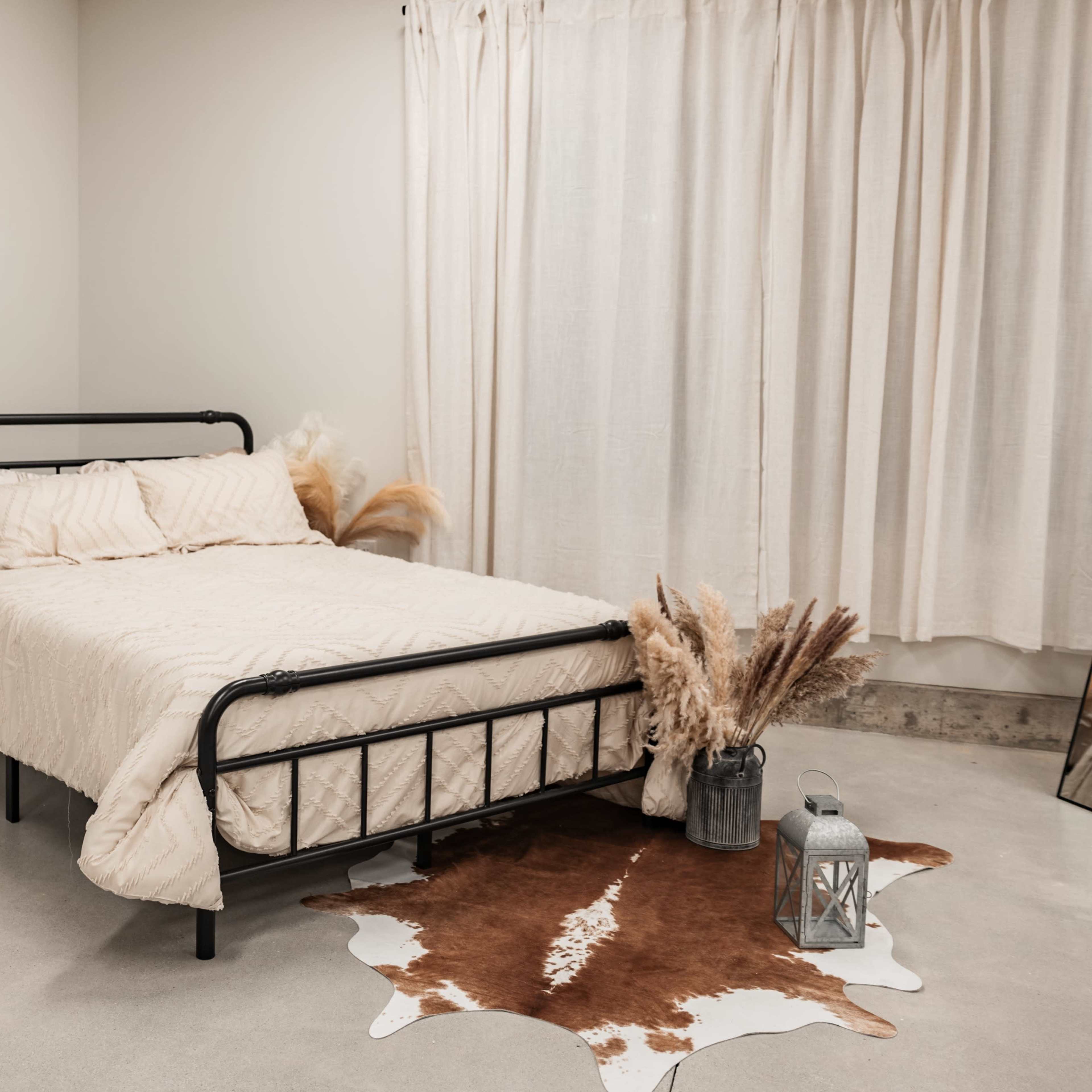 A metal bed with a beige quilt is positioned in a minimalist room featuring a decorative plant, a cowhide rug, and a lantern beside a large mirror.