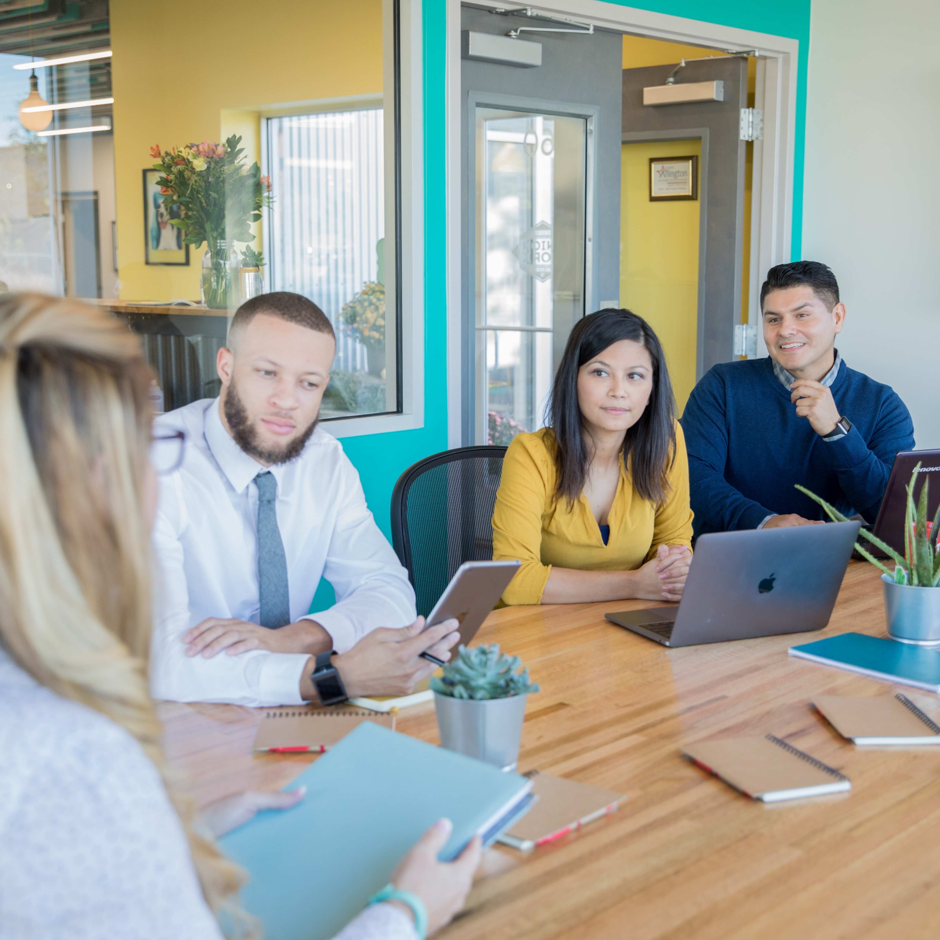 A group of four professionals engages in a meeting around a wooden table in a brightly colored office.