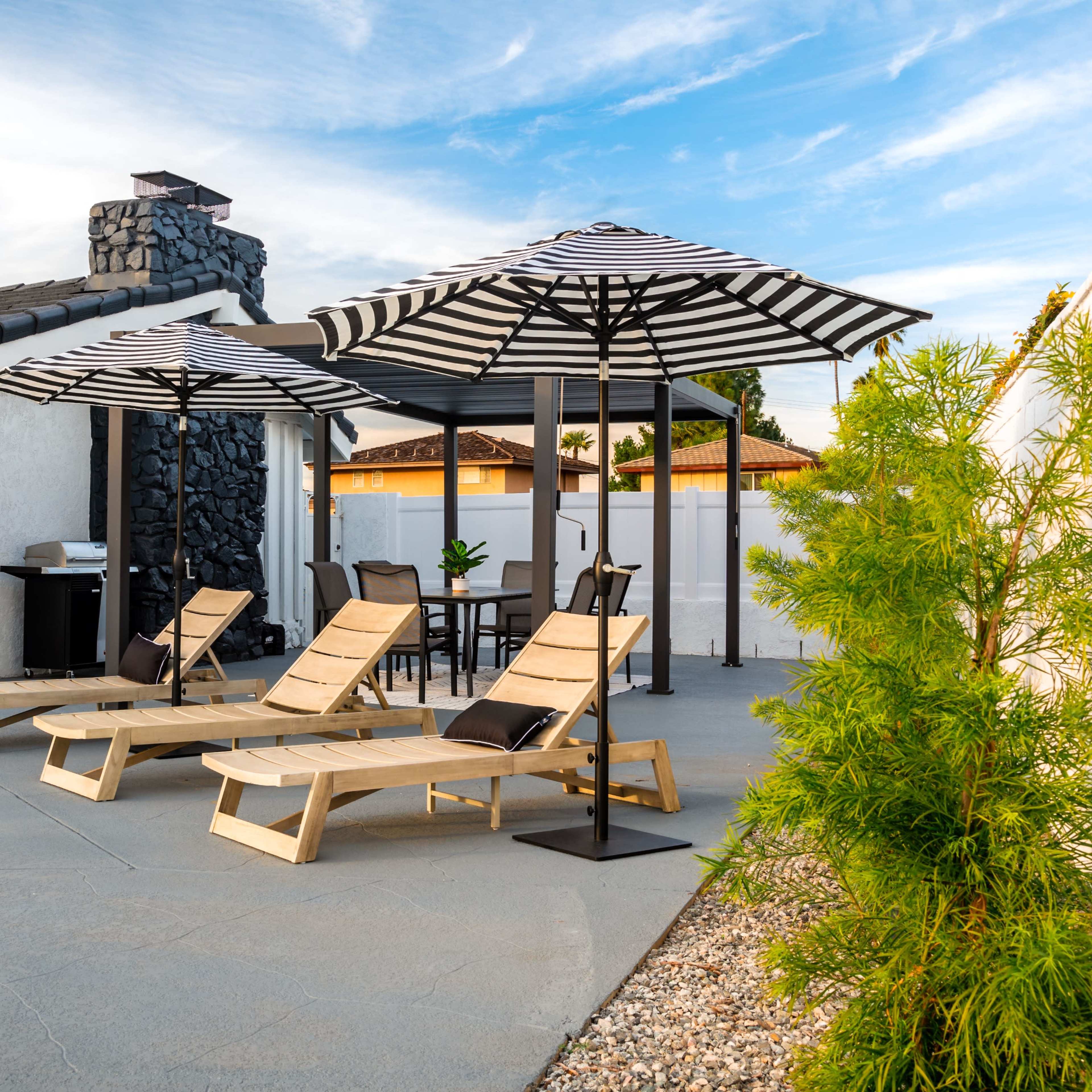 A backyard patio with two lounge chairs under striped umbrellas, a stone fireplace, and a grill in the background.