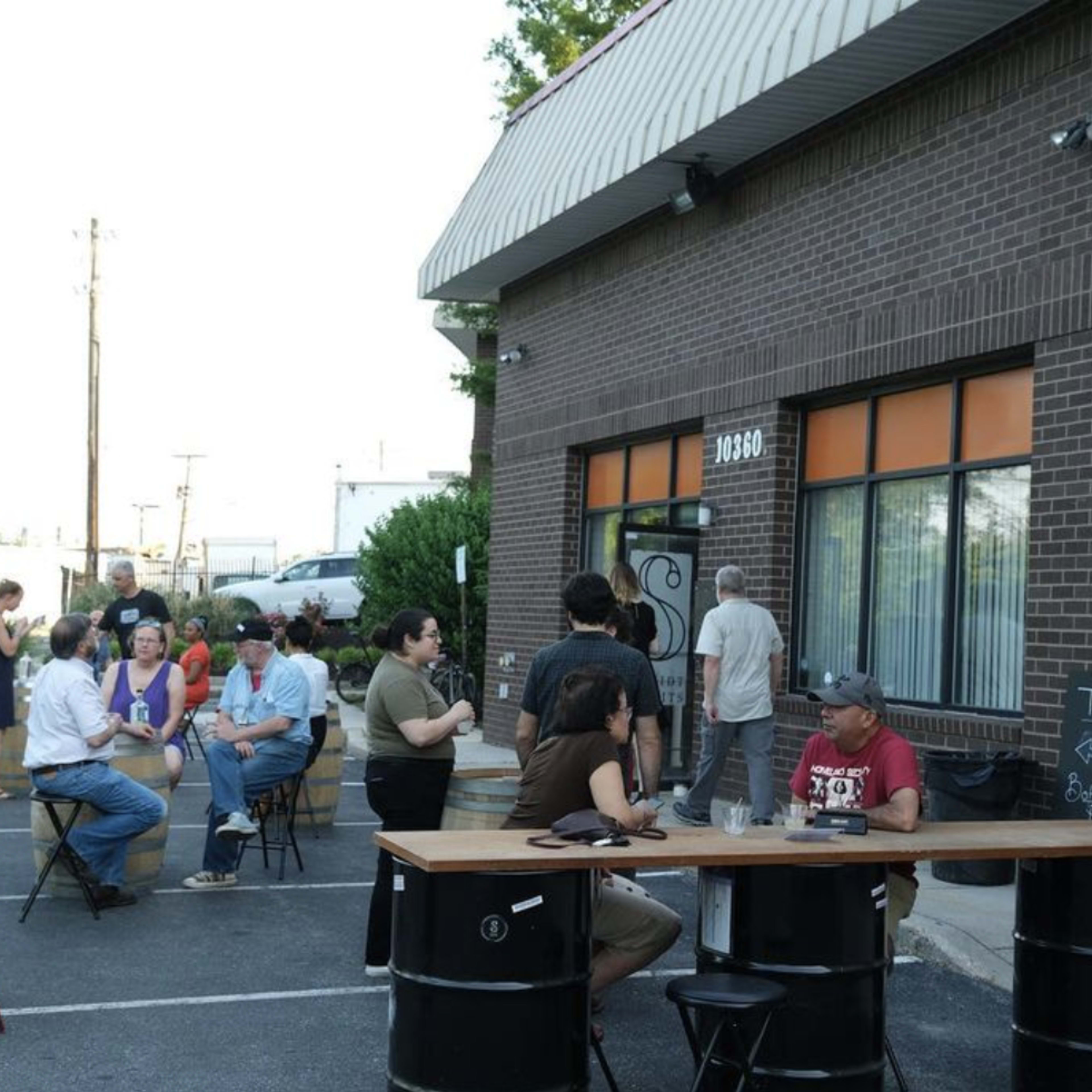 A group of people sits around outdoor tables in front of a distillery, with a large copper still visible nearby.