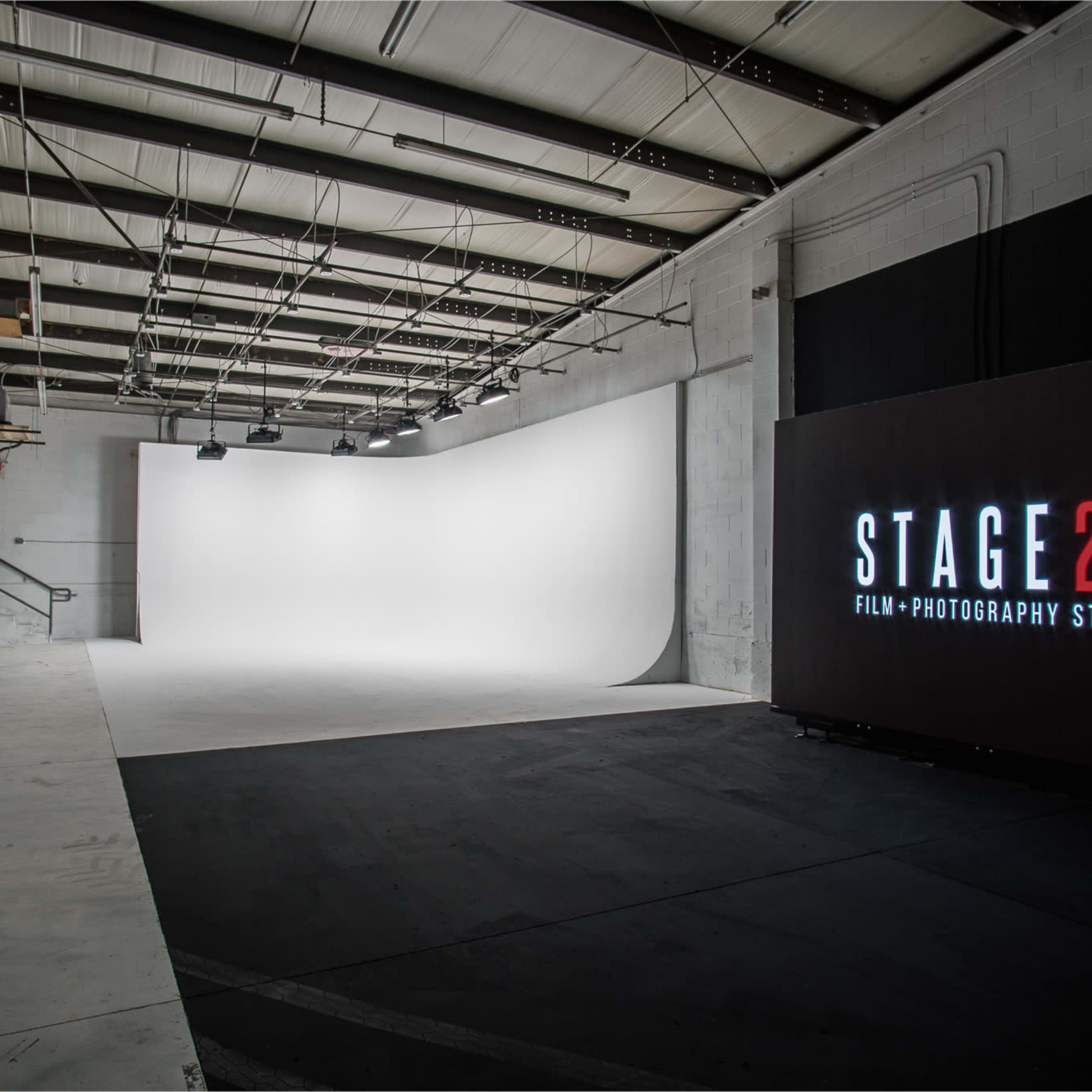 An empty photography studio featuring a white backdrop, black flooring, and a large illuminated sign reading "STAGE27 FILM + PHOTOGRAPHY STUDIO."