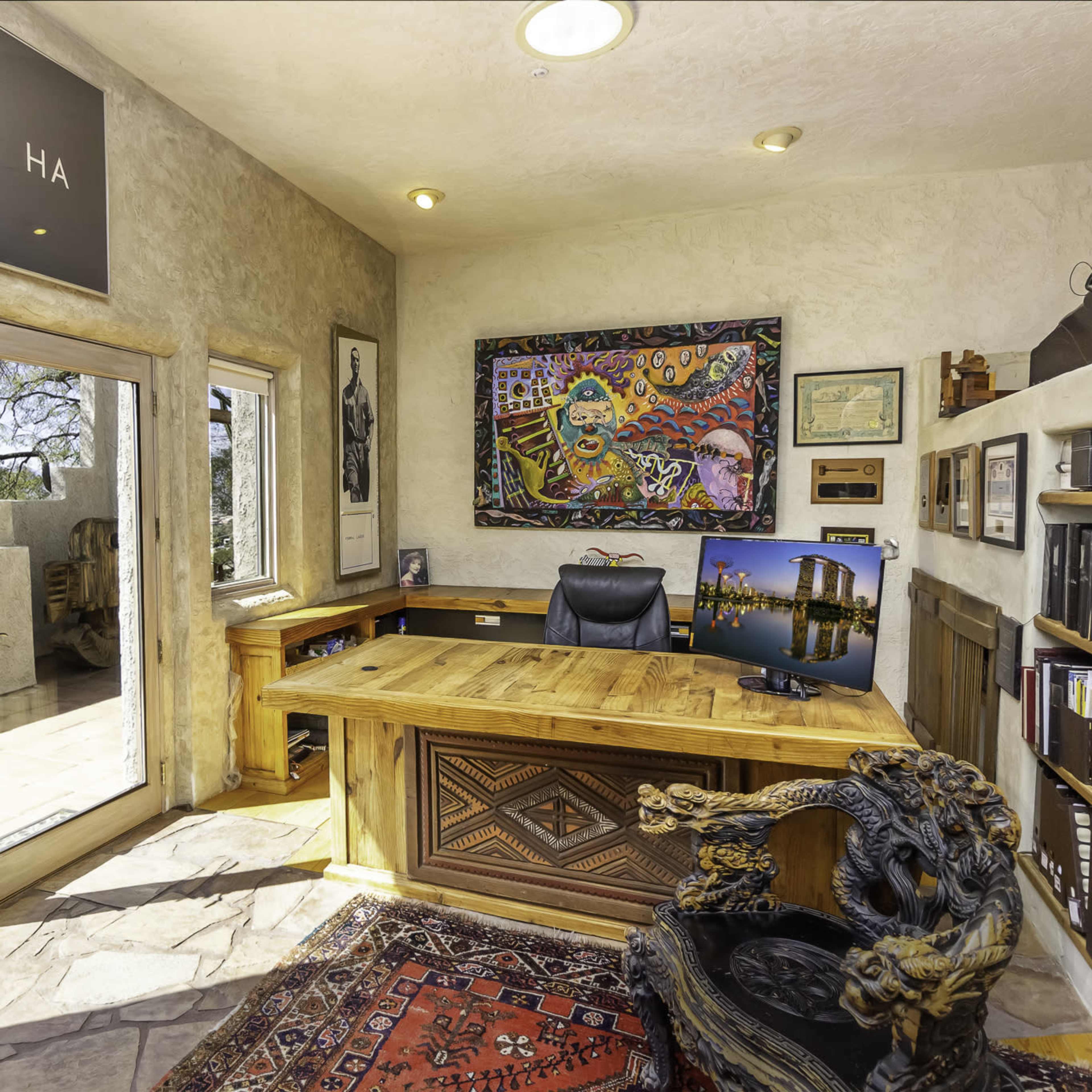 A well-furnished office with a wooden desk, a colorful artwork on the wall, and large windows overlooking a patio.