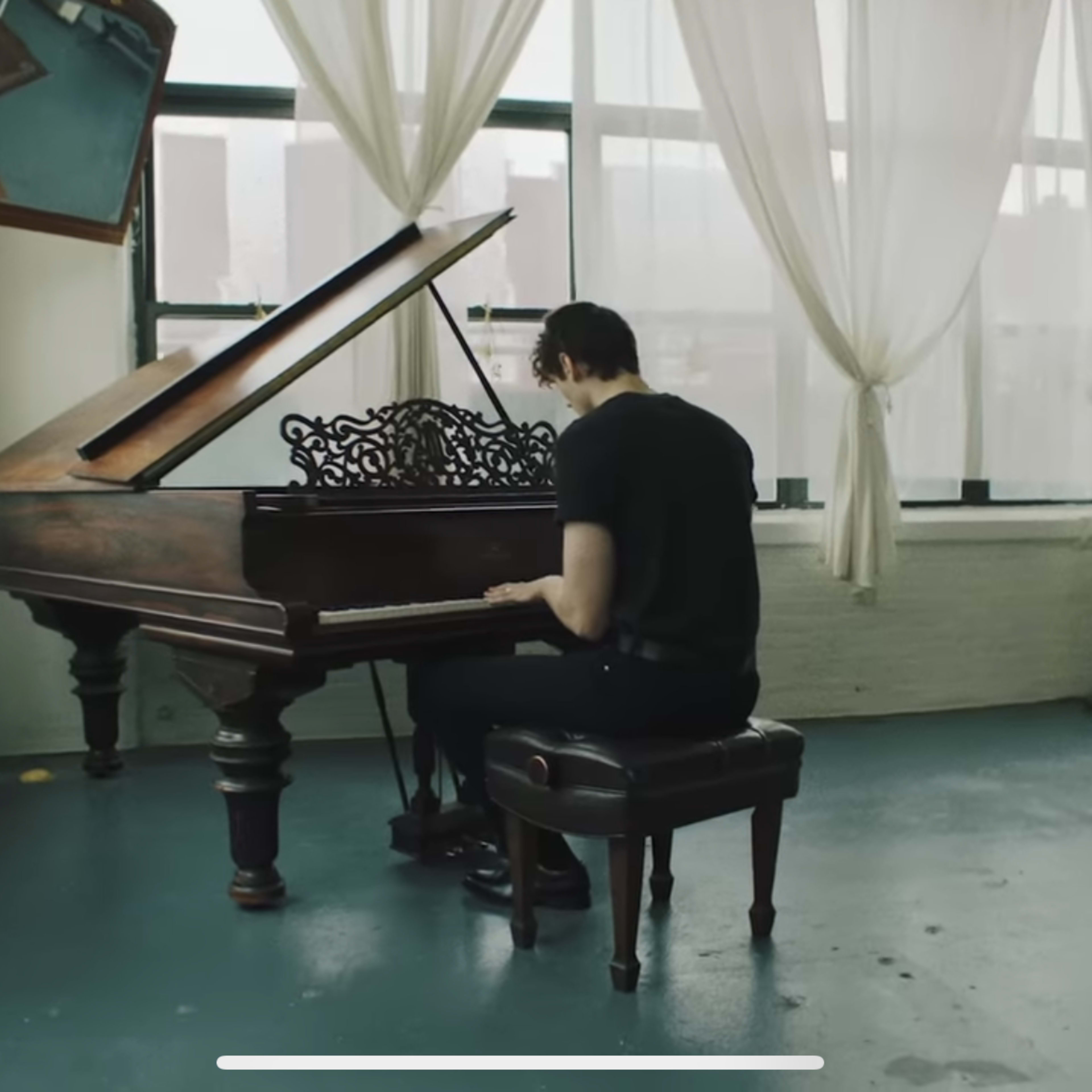 A person sits at a grand piano in a spacious, sunlit room with sheer curtains and an artwork mounted on the wall.