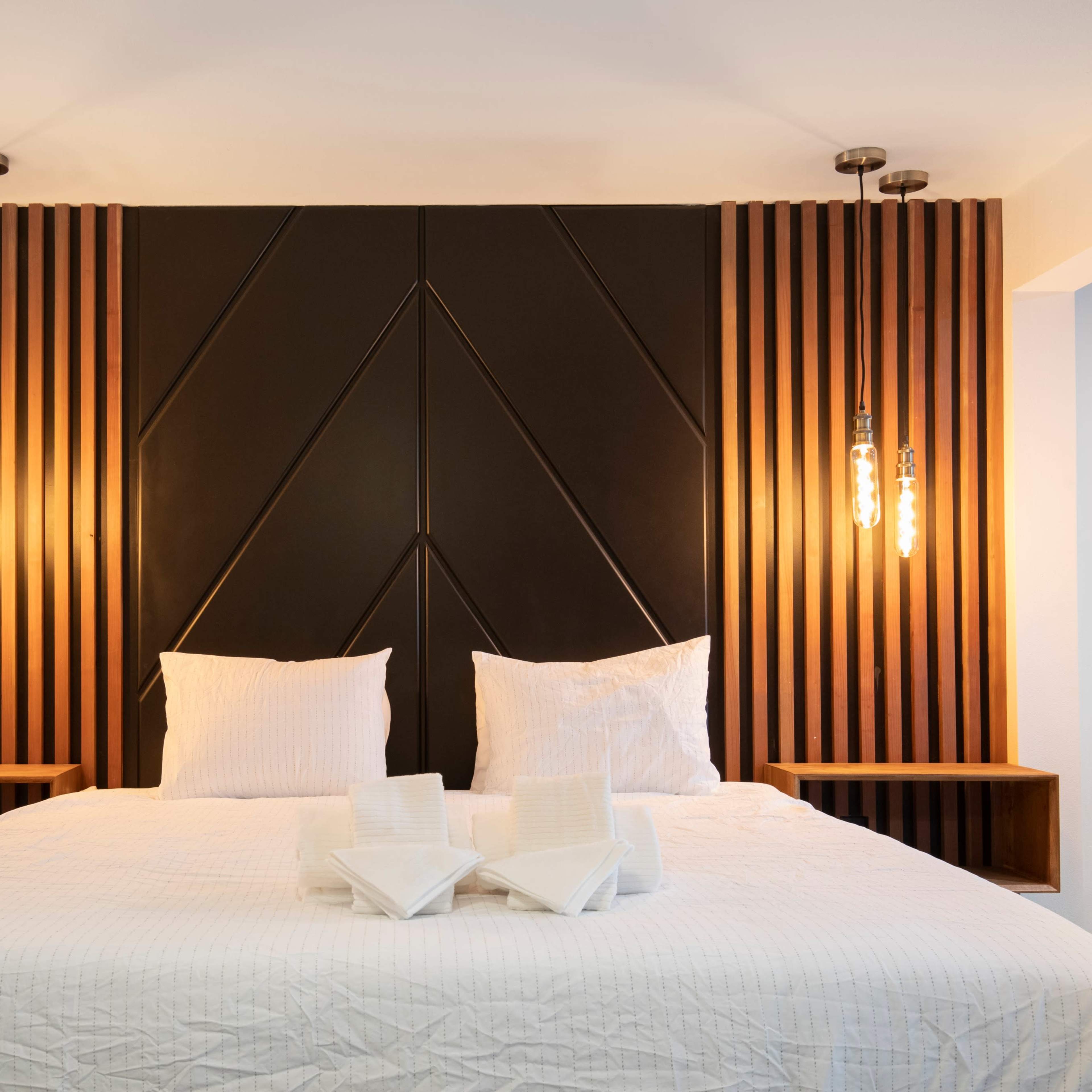 A modern bedroom featuring a bed with white bedding, a dark wood accent wall, and two pendant lights on either side.