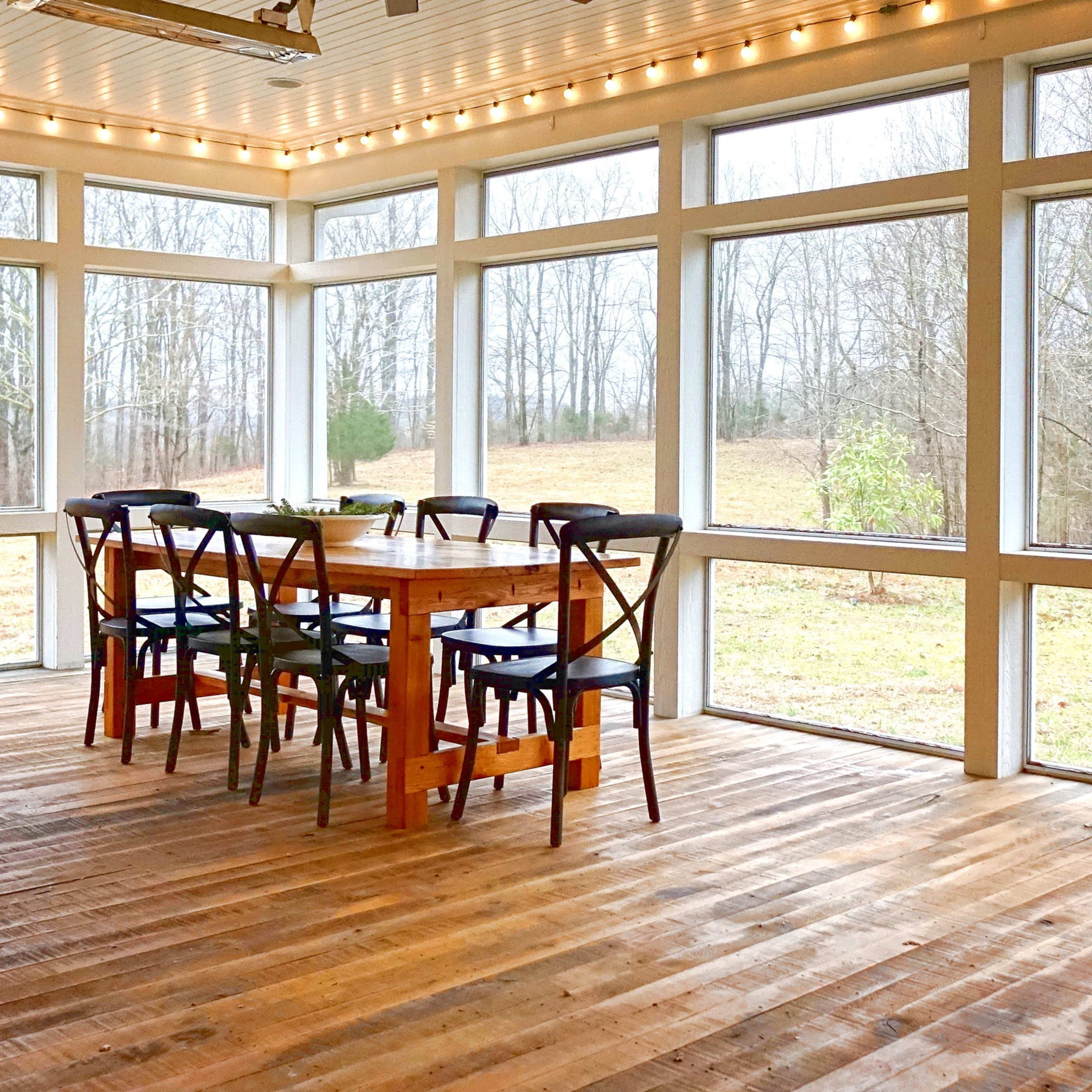 A spacious room features a wooden dining table surrounded by chairs, with large windows offering a view of a grassy area and trees outside.