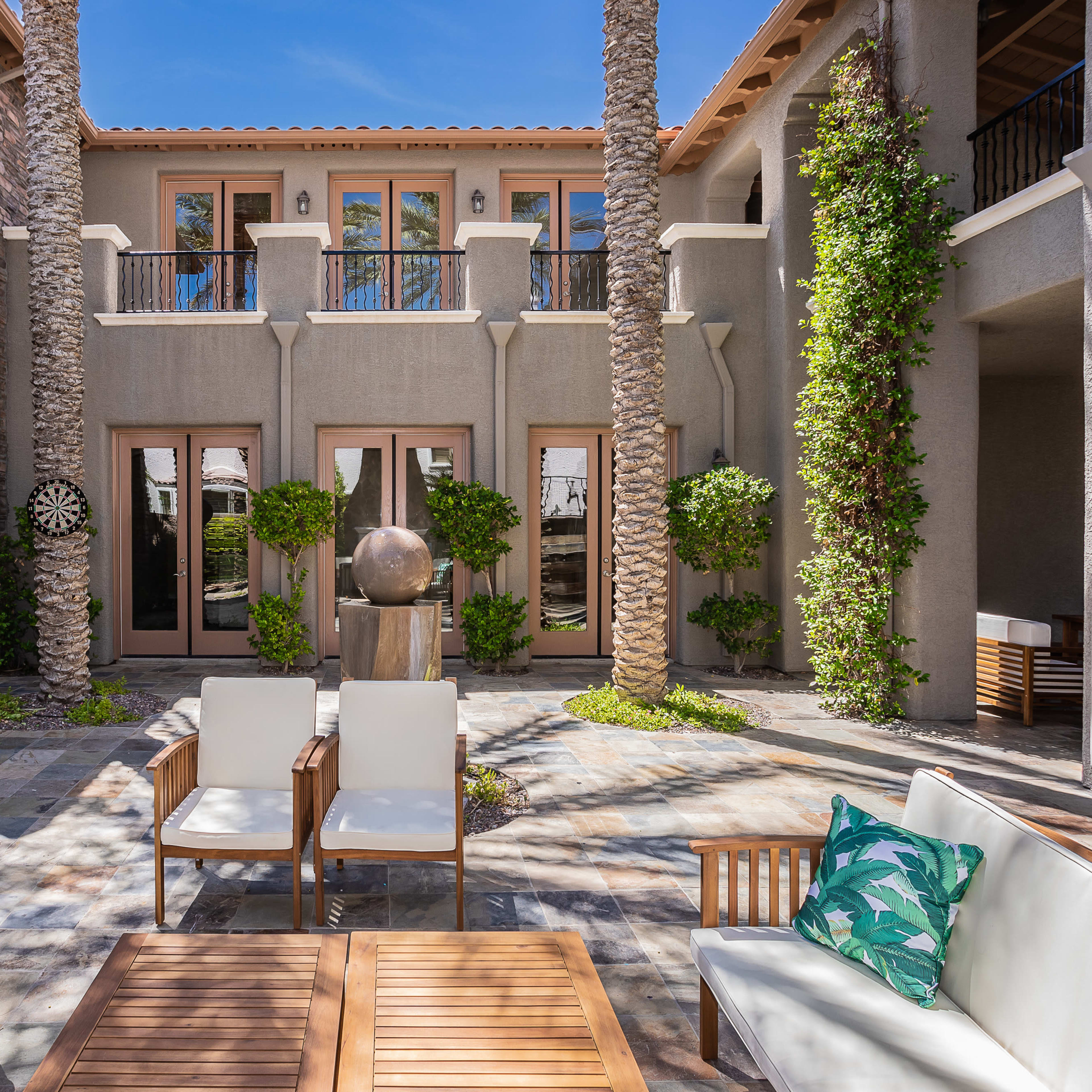 A landscaped courtyard featuring modern seating, palms, and a decorative sculpture, framed by two adjacent buildings.