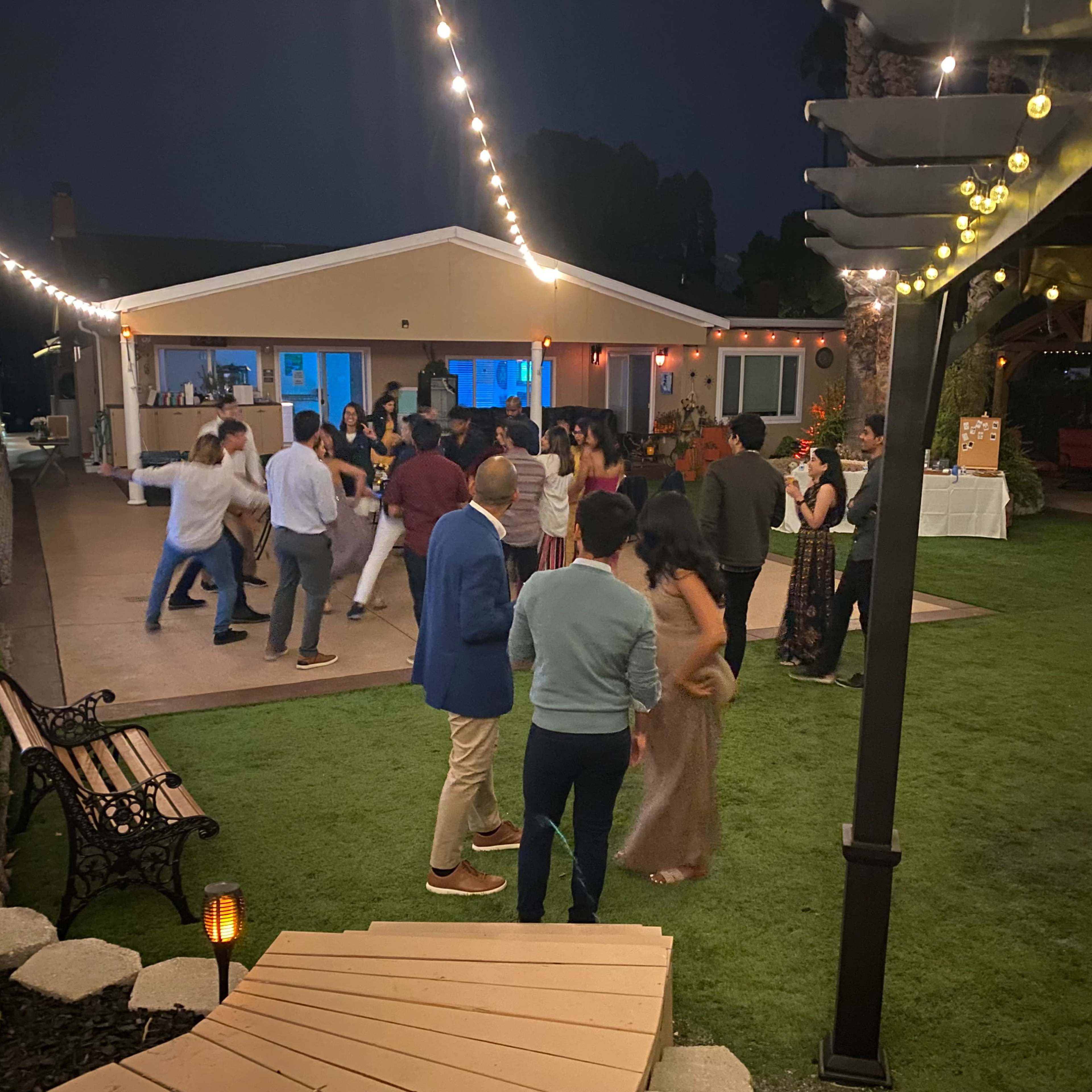 A lively outdoor event is taking place in a well-lit backyard with guests dancing, seated at benches, and enjoying the festivities near a house.