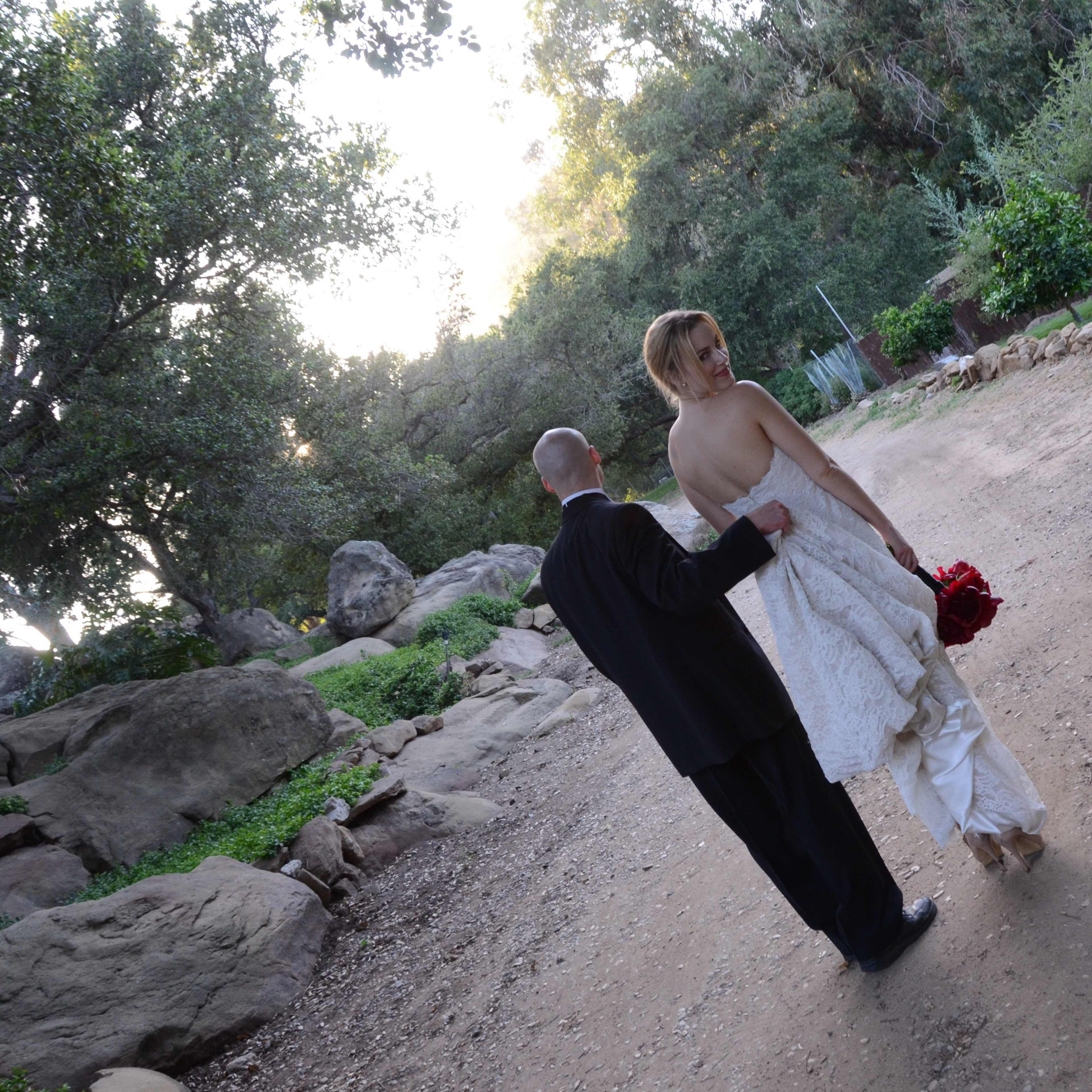 A bride in a white wedding dress holds a bouquet while walking away with a man in a black suit along a gravel pathway surrounded by trees and rocks.