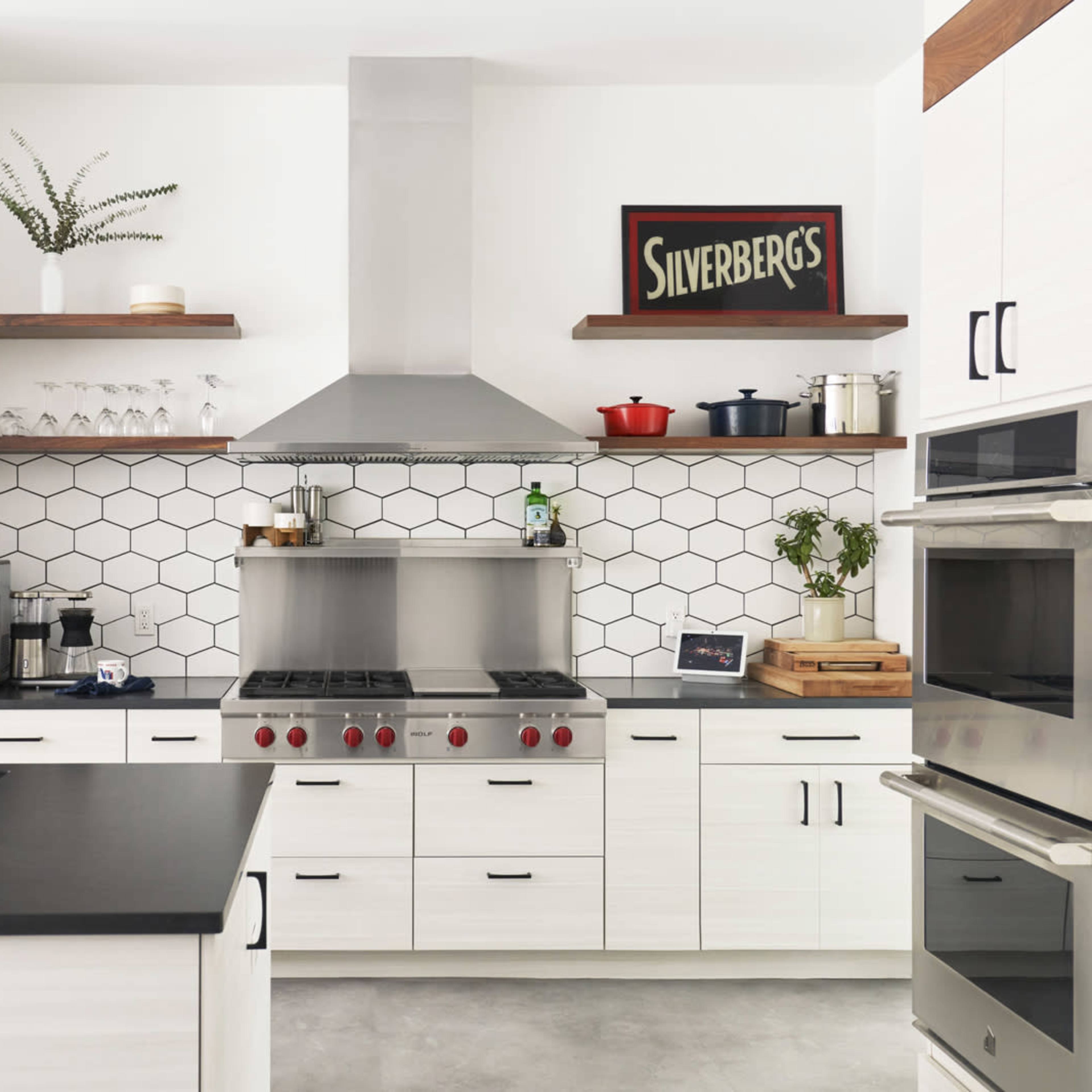 A modern kitchen features white cabinetry, a hexagonal tile backsplash, metal appliances, and a central cooking area with a gas range and overhead hood.