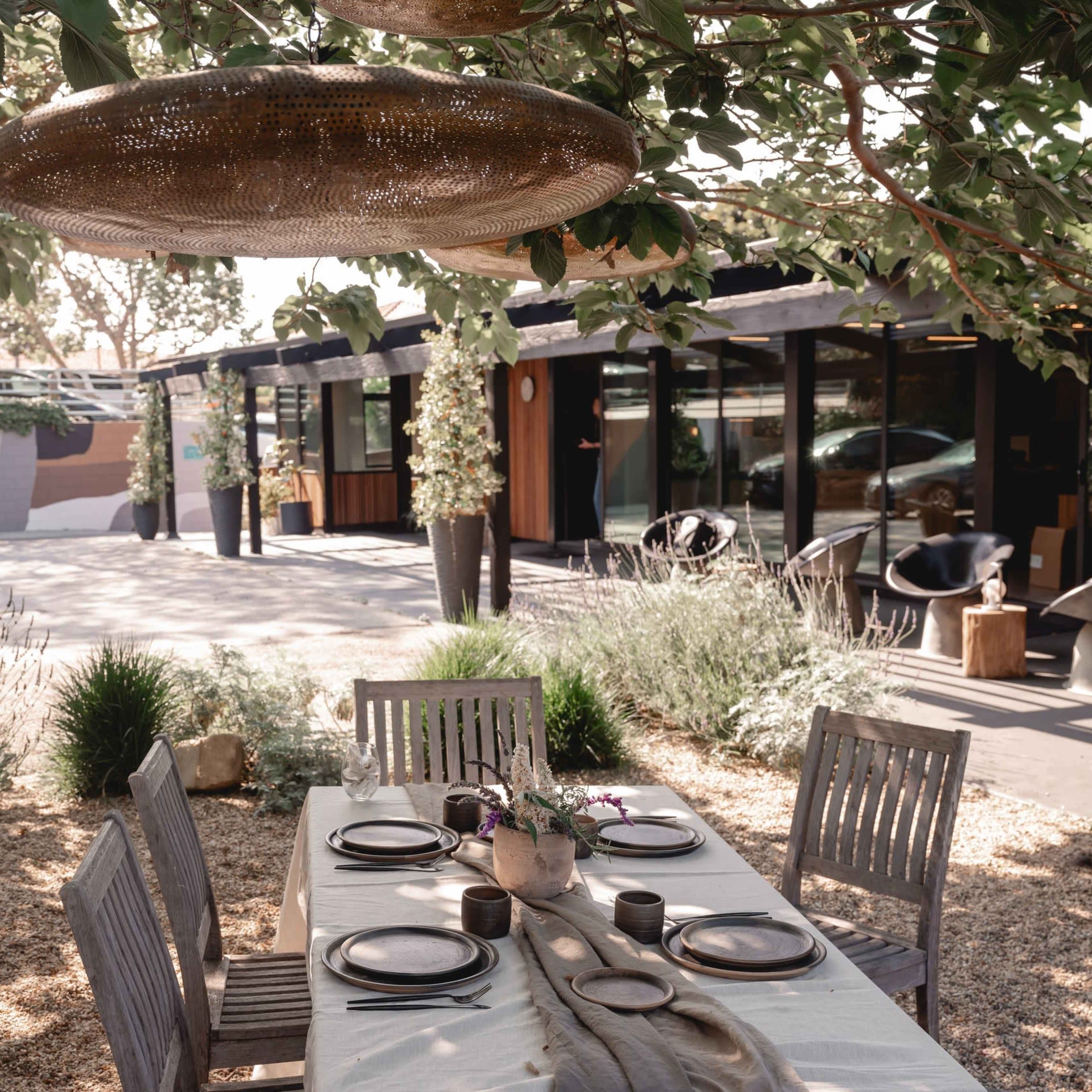 A wooden dining table is set with plates and decorative items under a tree in a patio area adjacent to a modern building.